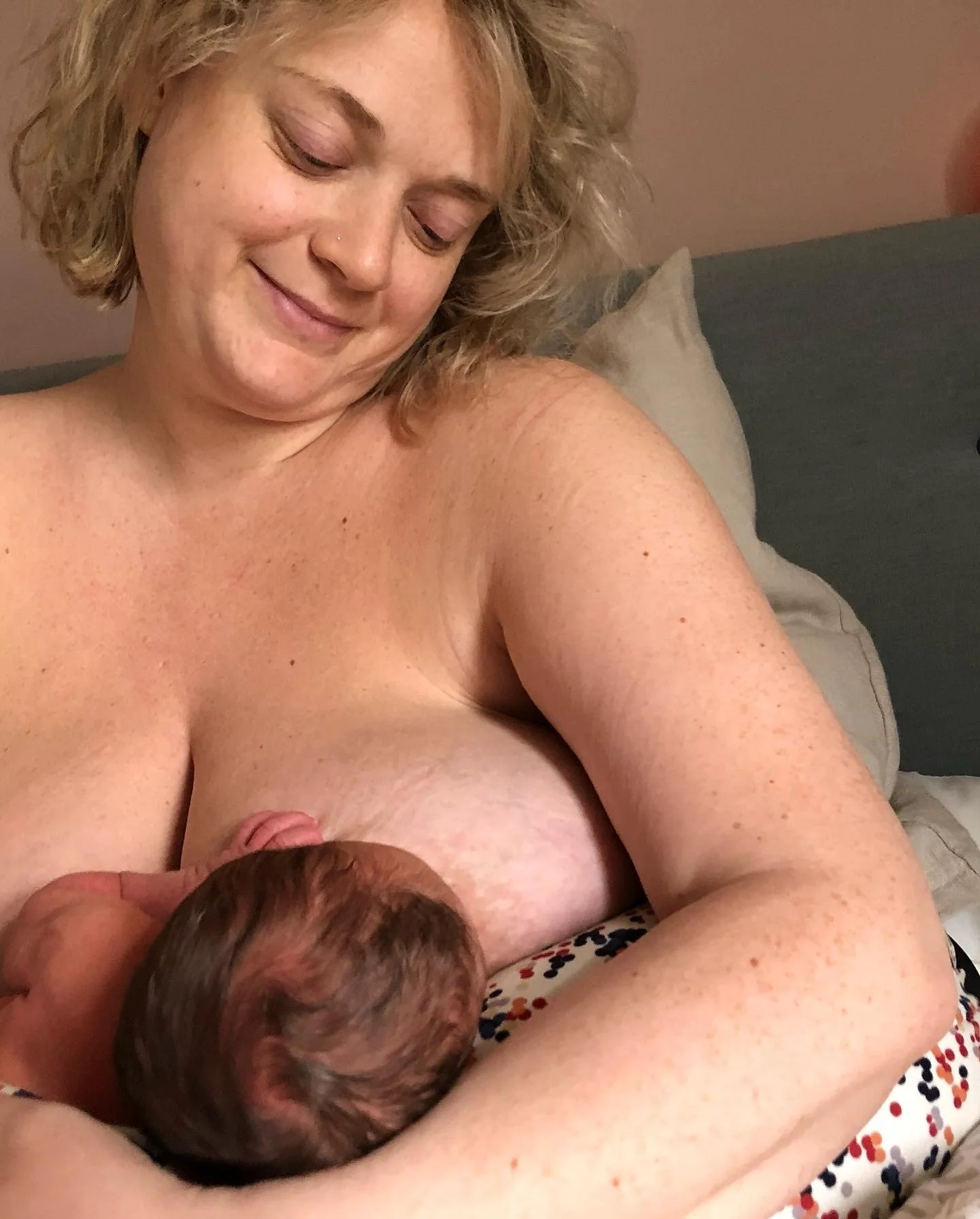 A woman with short curly blonde hair and a nose piercing breastfeeds a baby while sitting on a bed with a pillow in the background.