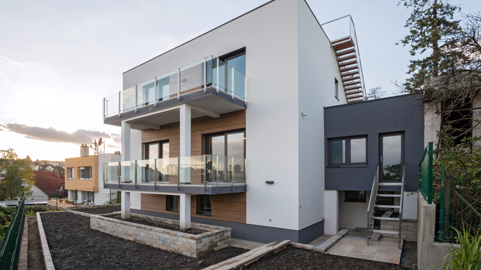Neues modernes Mehrfamilienhaus mit weißen und dunklen Fassaden, großen Balkonen mit Glasgeländern, Außenaufgang, Gartenbereich und angrenzendem Nachbarhaus.