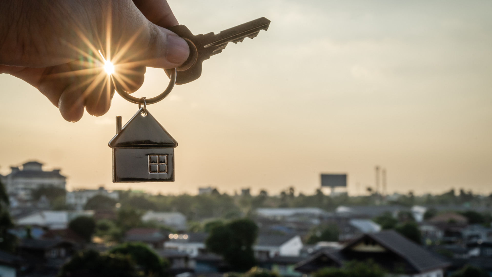 Hand hält einen Schlüssel, an dem eine Haus-Schlüsselanhänger hängt, vor einem Sonnenuntergang über einer Stadt.