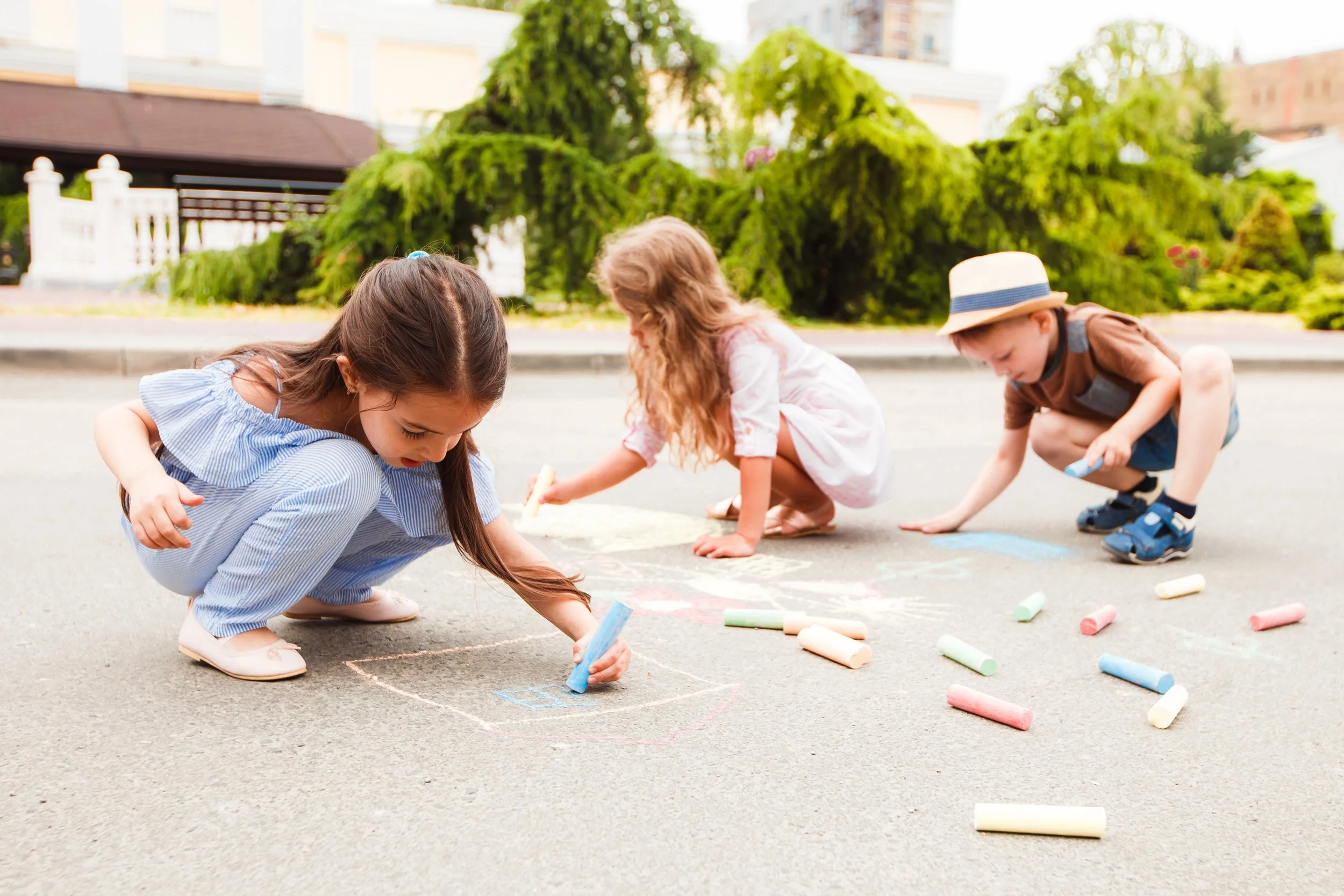 Drei Kinder zeichnen mit bunten Kreide auf die Straße, umgeben von Kreide und Hüpfen, im Hintergrund Bäume und Häuser.