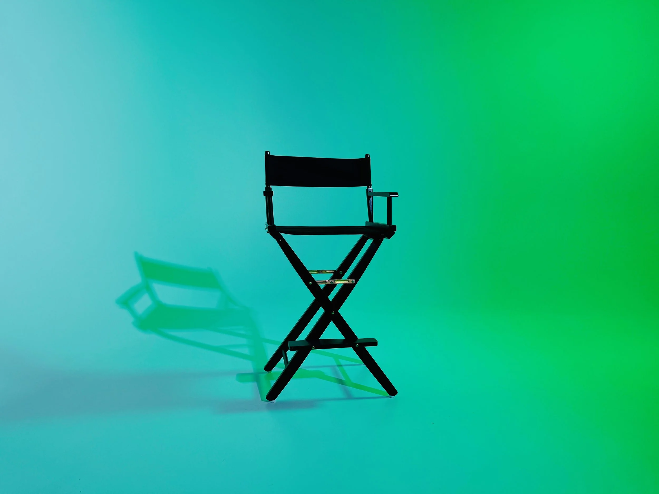 A black director's chair against a colorful gradient background in shades of green, blue, and yellow, casting a shadow on the surface.