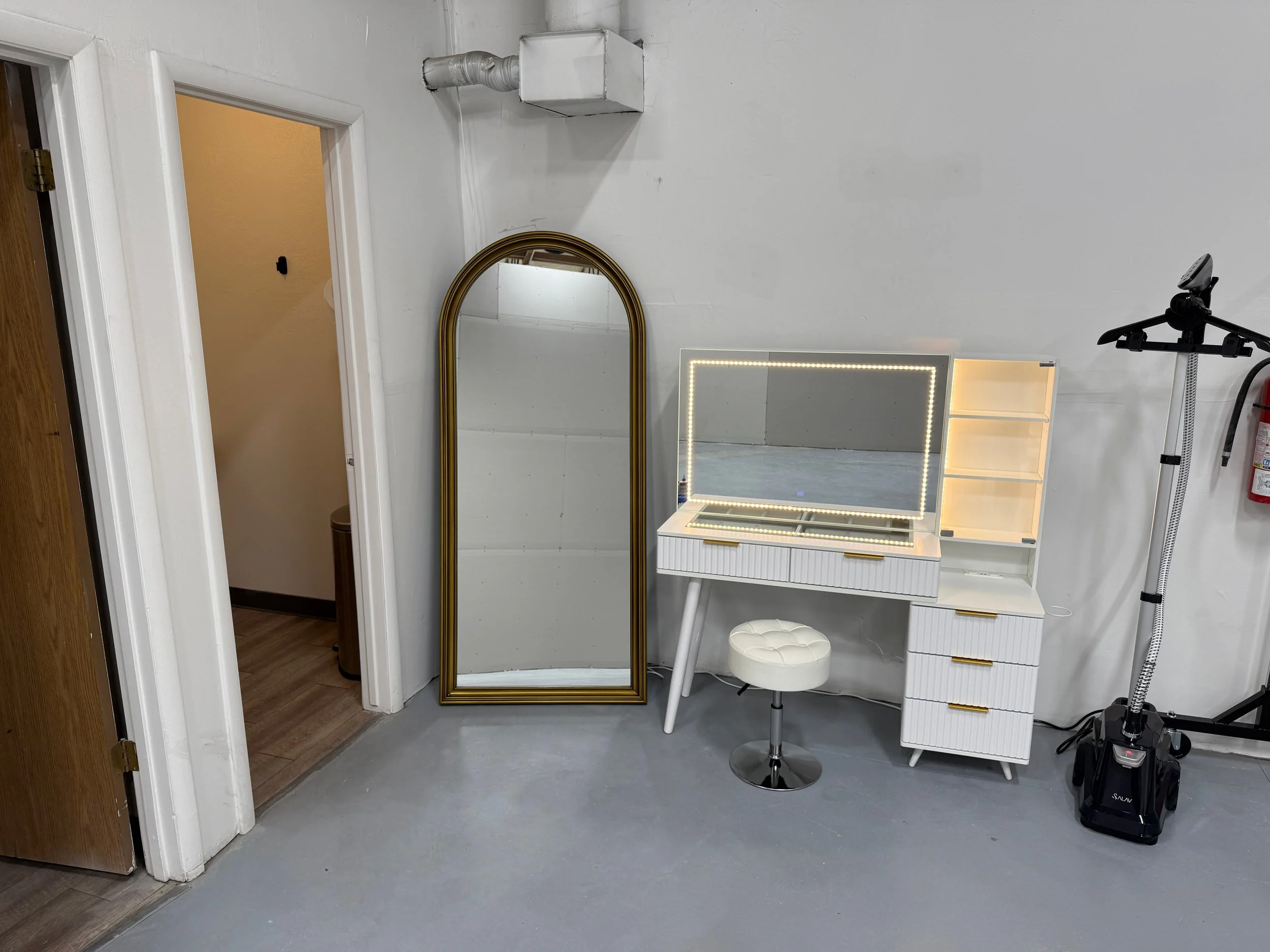 A room with a white wall, a mirror with a gold frame, a vanity with a mirror LED light, a small white stool, a small white cabinet, a black and silver vacuum cleaner, and an exercise machine with a screen and a handle.