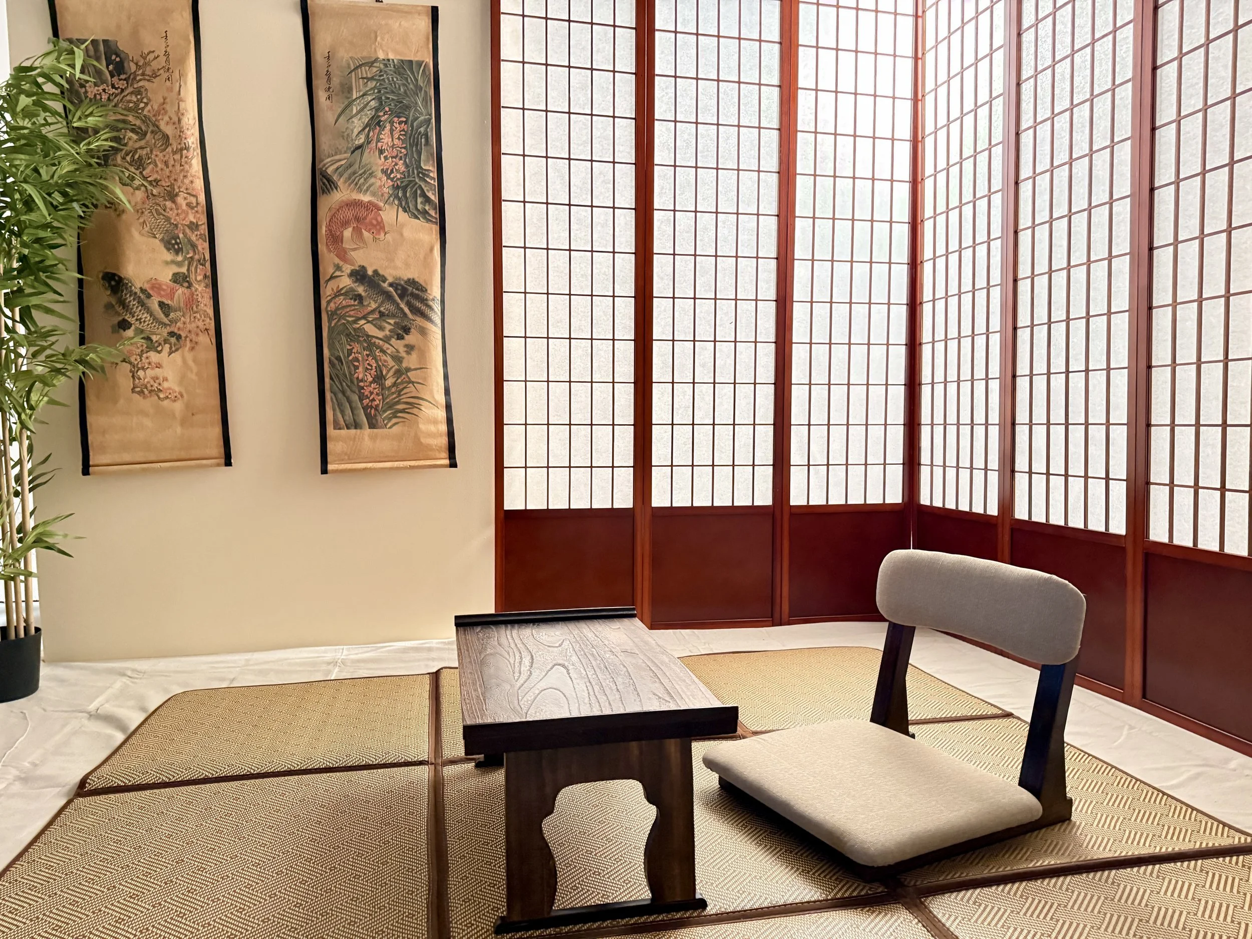 A Japanese-style room featuring a shoji screen window, two hanging traditional artwork scrolls with nature and fish scenes, a potted bamboo plant, a dark wooden table, and chairs with light cushions.