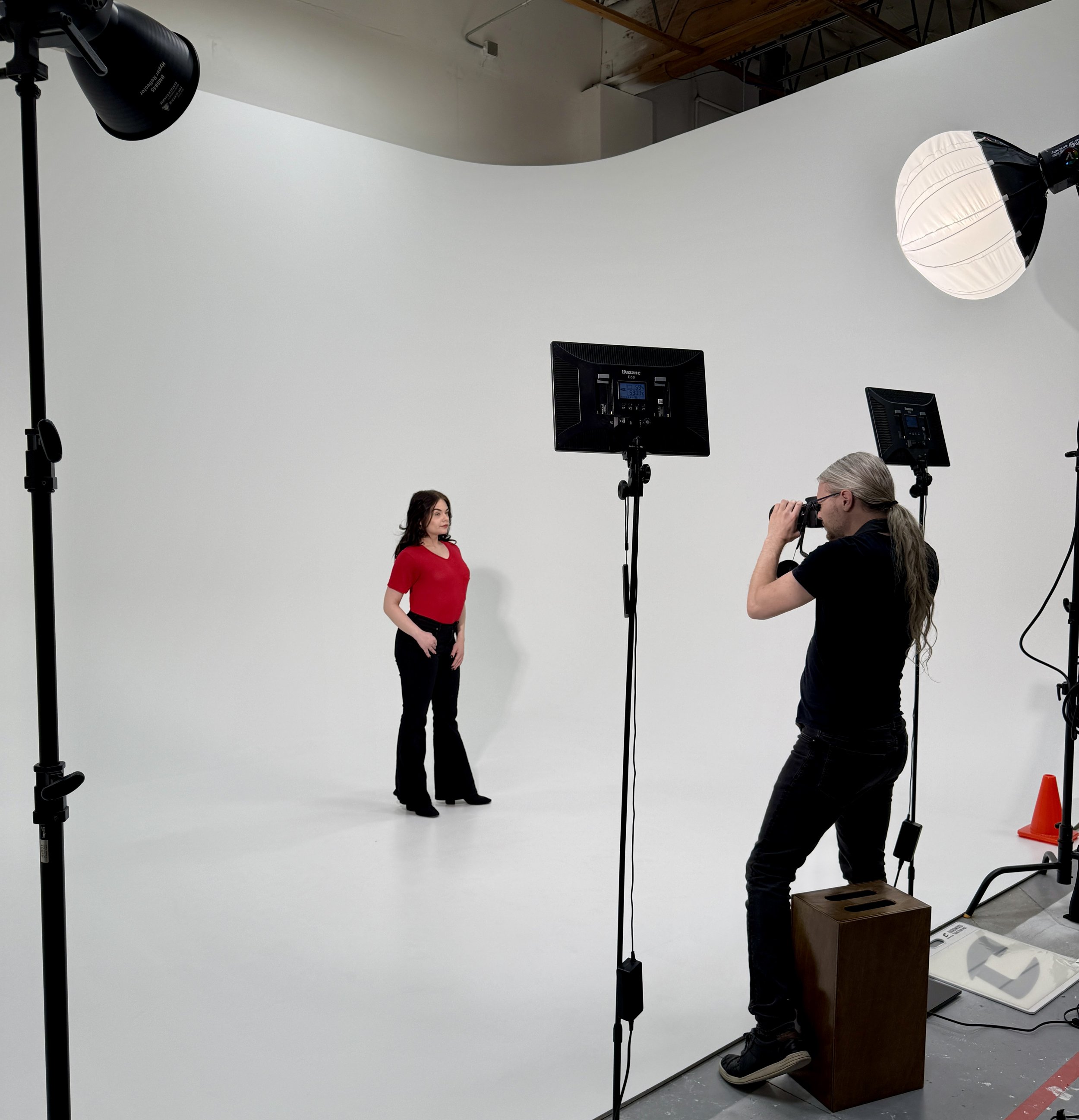 A photoshoot studio setup with a female model in a red shirt and black pants standing in front of a white backdrop, with a photographer taking her picture. Studio lights, reflectors, and equipment are visible.
