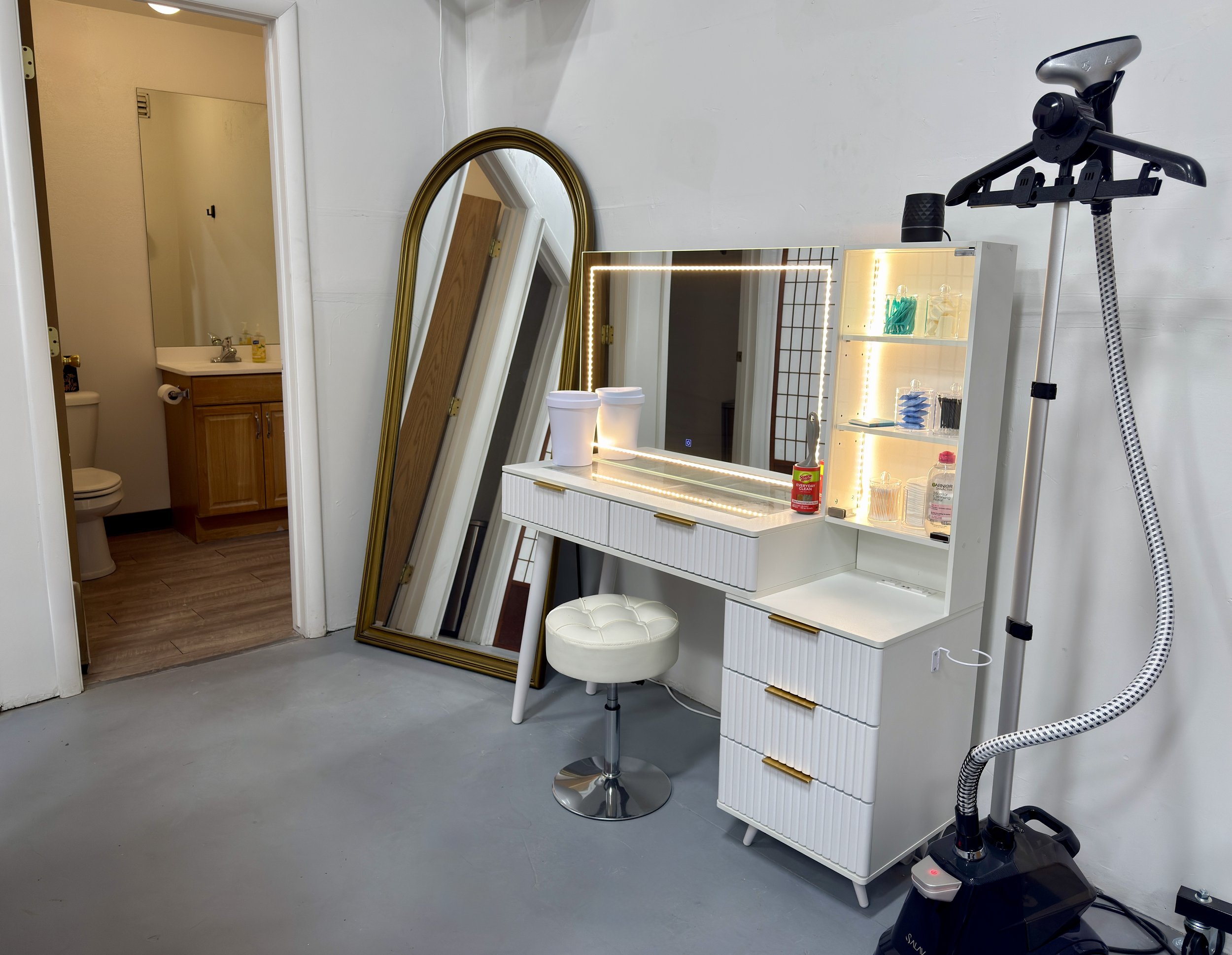 A room with a white vanity mirror with built-in lights, a white stool with a cushioned seat, a white shelving unit with bottles, jars, and makeup tools, a mirror leaning against a wall, a gold-framed mirror, a small bathroom with a wooden cabinet, a 