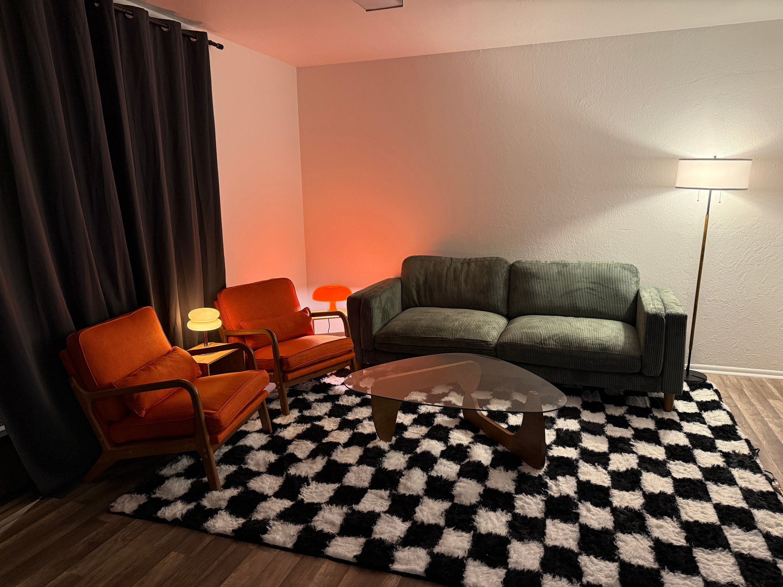 Living room with black and white patterned rug, three orange chairs, a glass coffee table, a green couch, a floor lamp, and a window with dark curtains. The room has white walls and wood flooring.