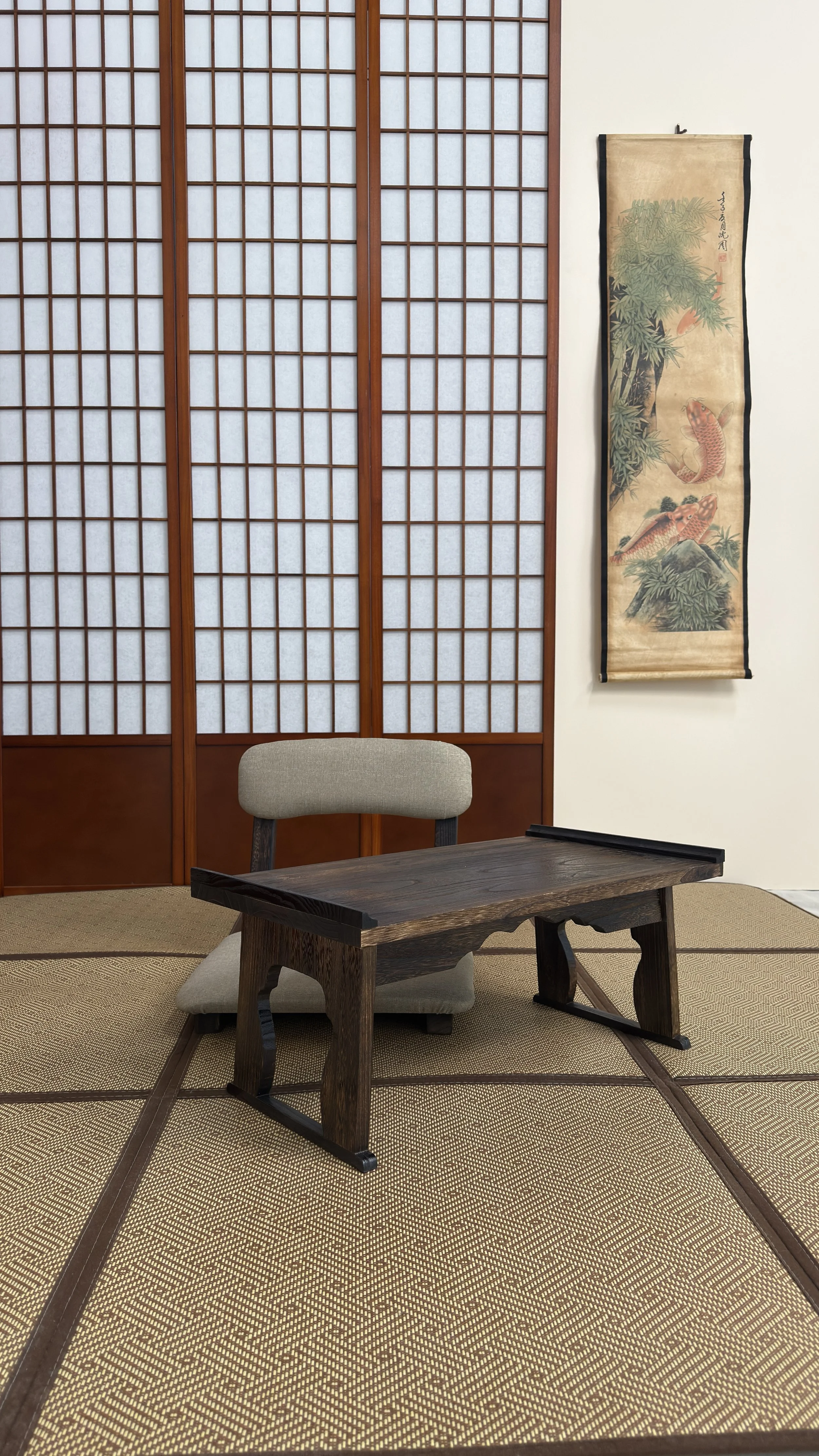 Interior of a traditional Japanese room featuring shoji screens, a low wooden table, a Japanese-style chair, and a hanging scroll with artwork of koi fish and bamboo.