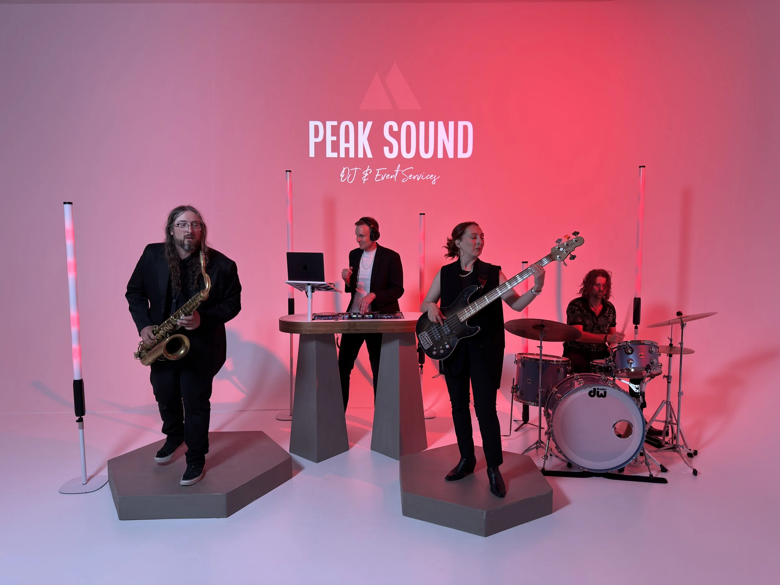 A band performing with four members on a pink gradient background. One person plays saxophone, another mixes on a laptop DJ setup, a third is playing bass guitar, and a fourth plays drums. The band is on a raised platform.
