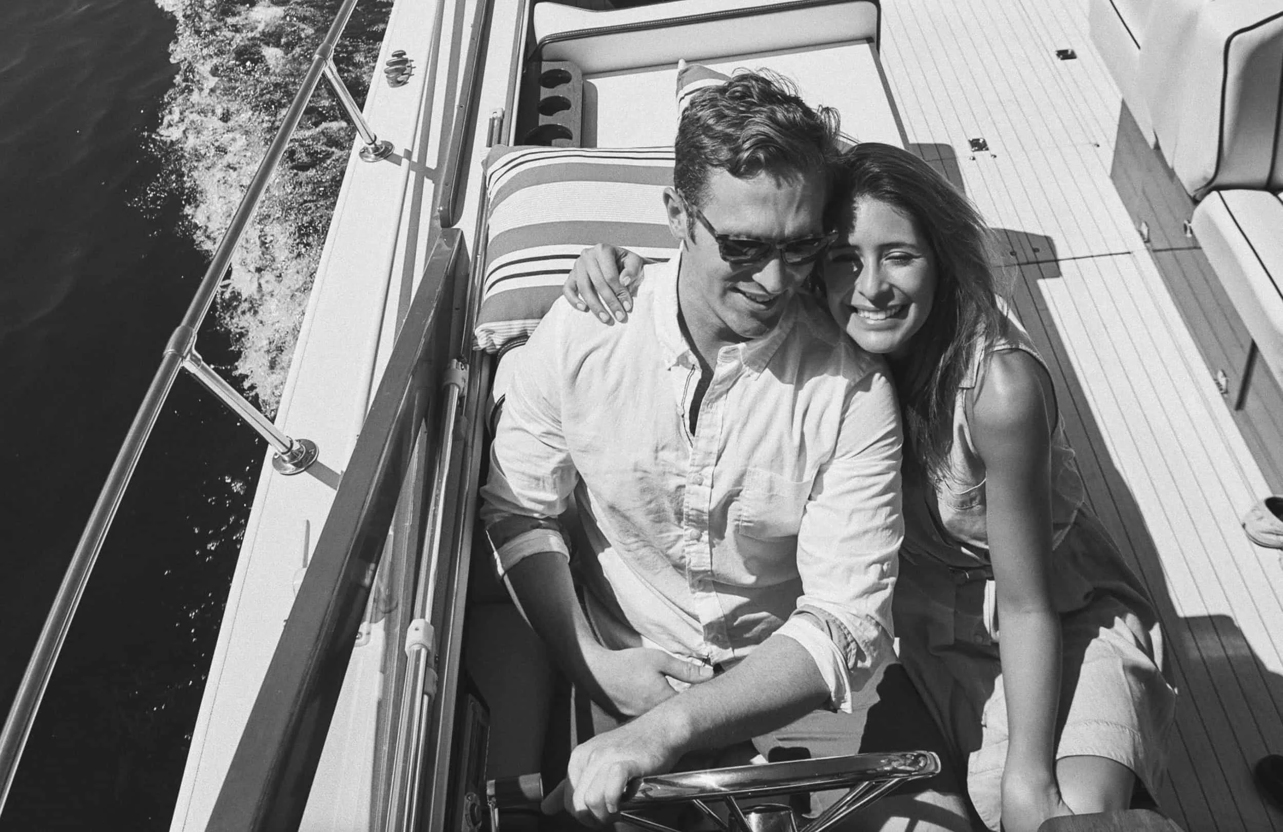 A couple smiling while sitting on a boat, with the man steering, in black and white.