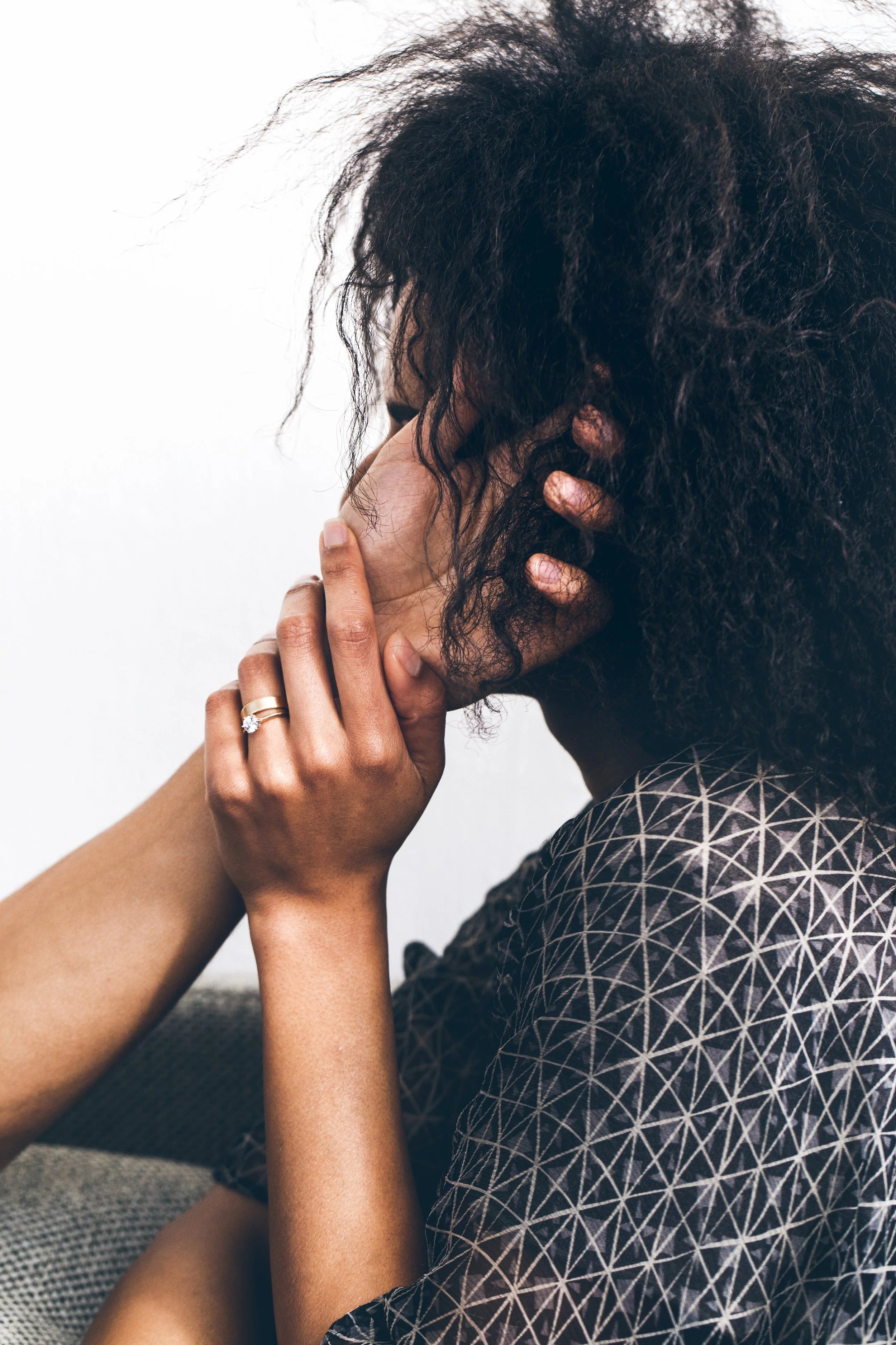 A woman with curly dark hair holding her face with her hand, wearing a ring, seated against a plain background.