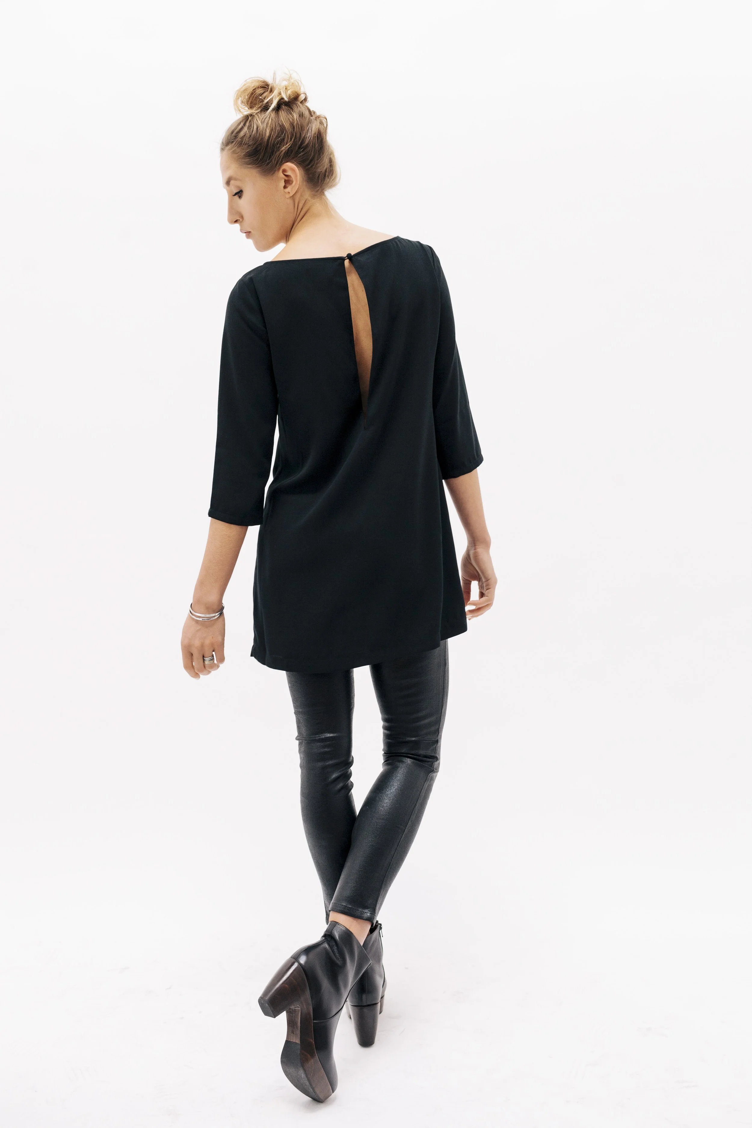 A woman posing with her back to the camera, wearing a black tunic with an open back detail, leather pants, and high-heeled ankle boots.