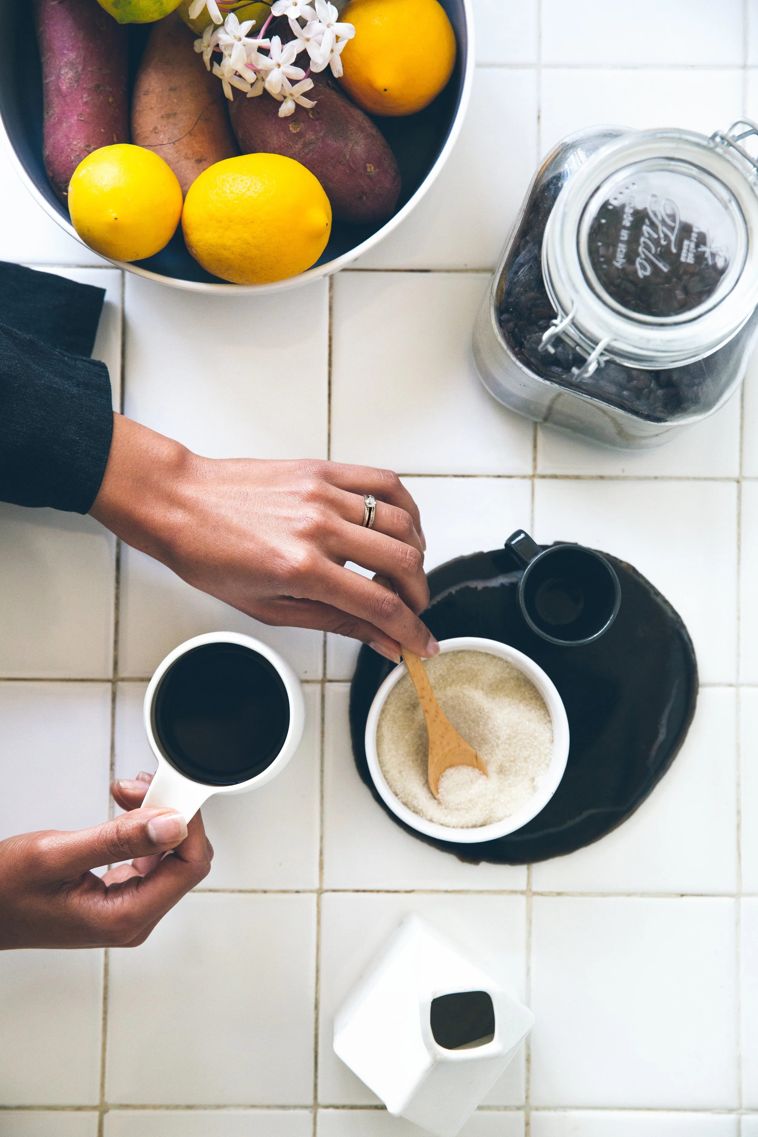 A person pouring coffee into a cup, with a bowl of sugar, a cup of coffee, and a jar of coffee beans on a white tiled countertop, along with a bowl of fruit and flowers.