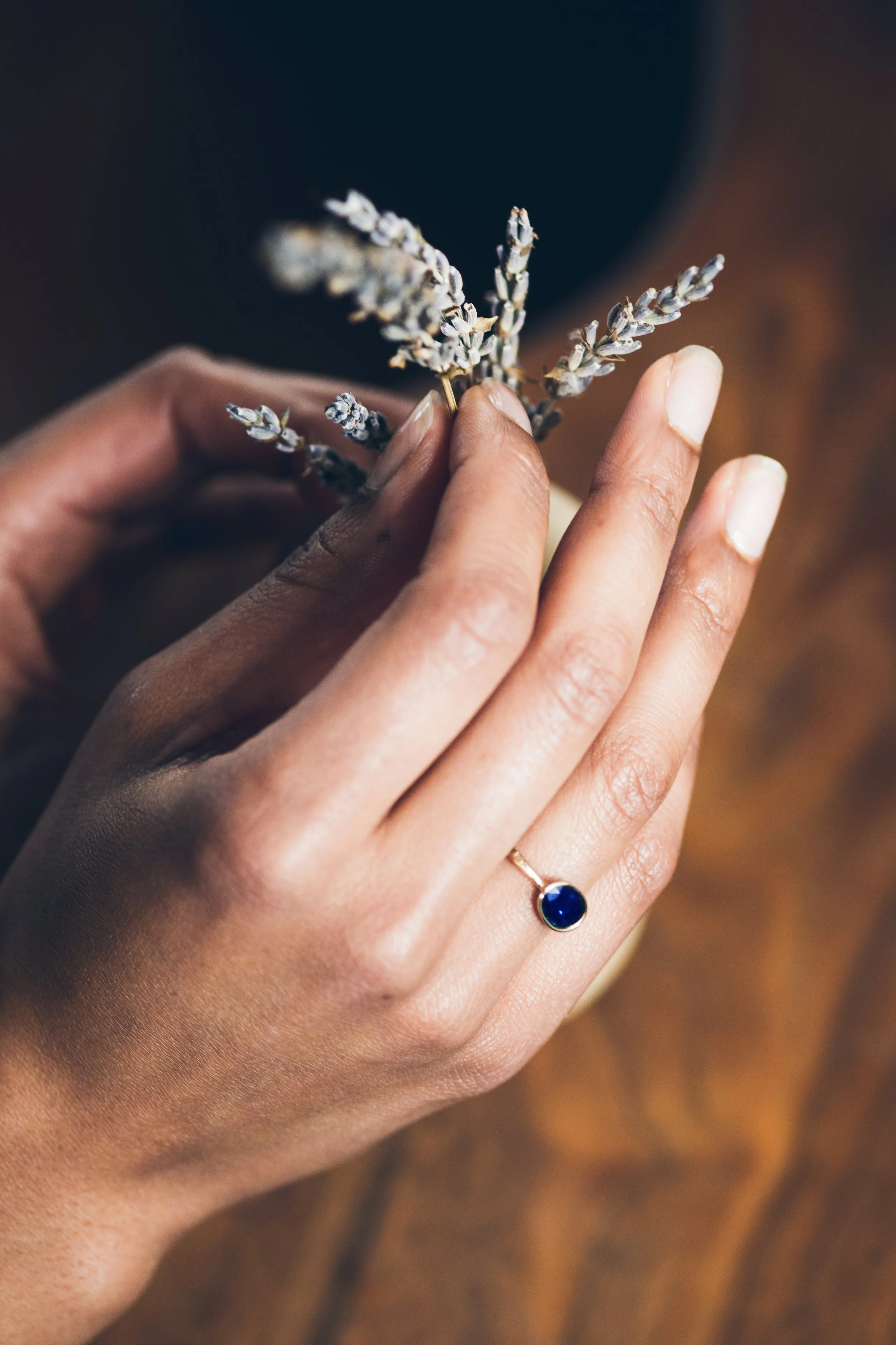 A person's hand with a gold ring featuring a large blue gemstone, holding a small bunch of dried lavender flowers.