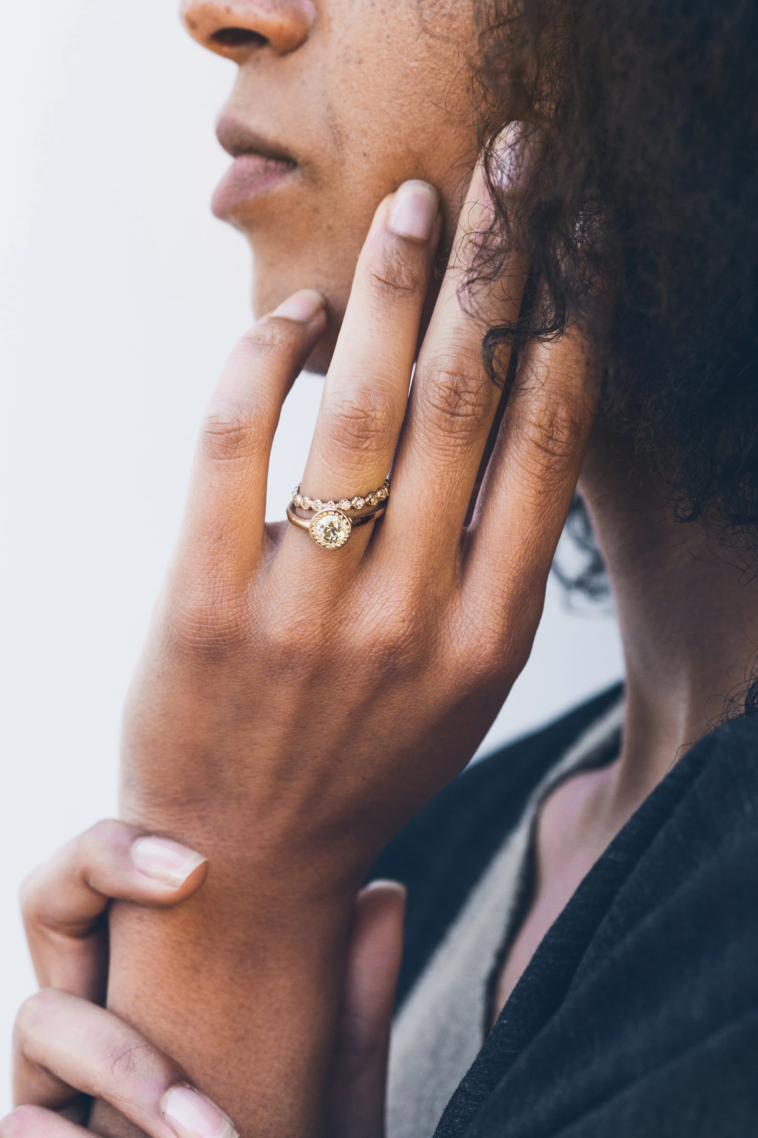Close-up of a woman with medium skin tone wearing rings, touching her face with her right hand. She has curly hair and is wearing a dark-colored garment.