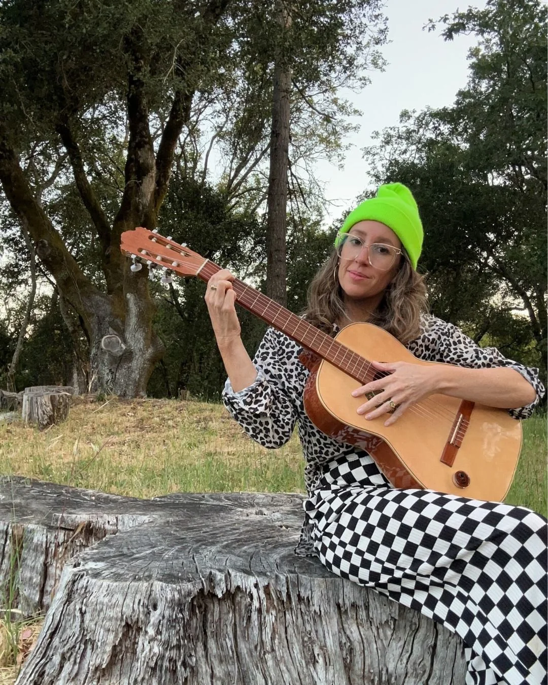 A woman taking a selfie in a mirror, making a peace sign with one hand, standing in front of a piano on a wooden parquet floor. She is wearing a yellow beanie, glasses, and a dark plaid shirt.