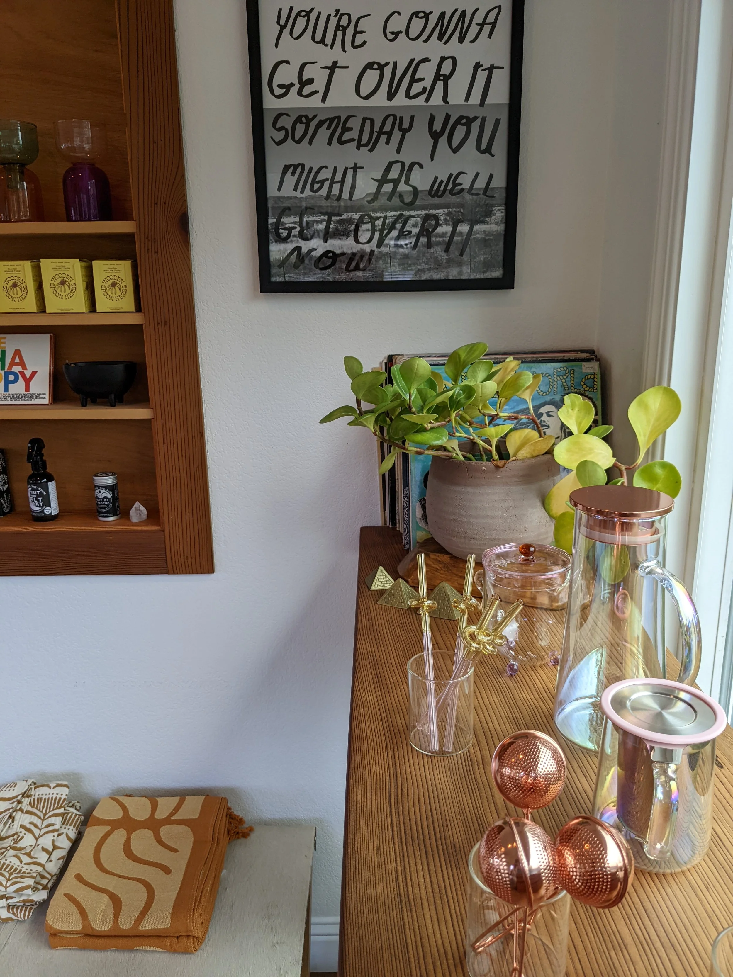 Decorative inside scene with a wooden table holding glassware and plants, framed quote on the wall, and shelves with small items and towels.