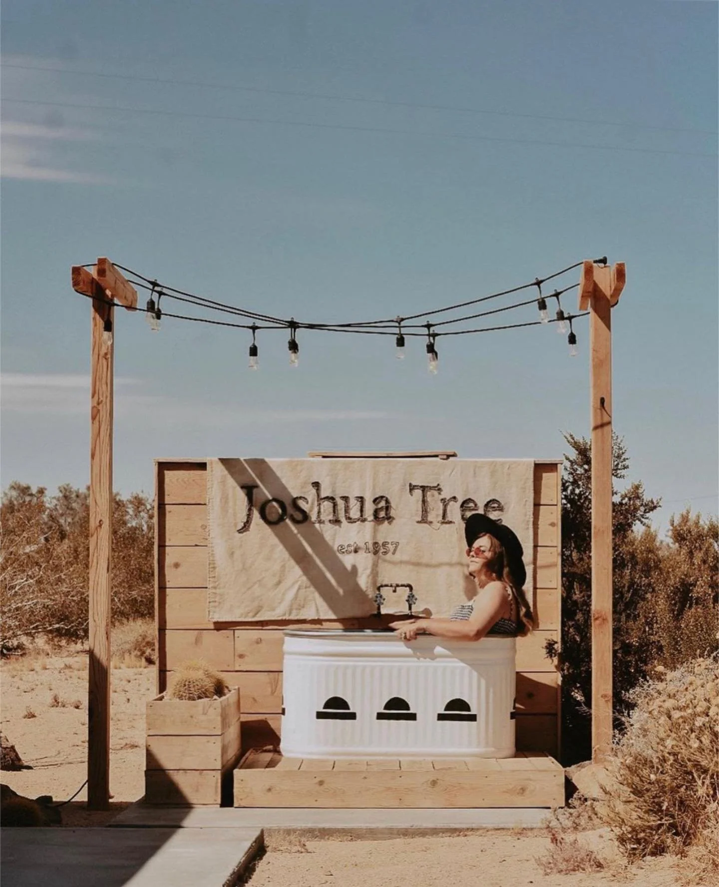 A woman sitting in a bathtub outdoors with a wooden sign behind her that reads "Joshua Tree est. 1957". She is wearing sunglasses and a black hat. The setting appears to be in a desert with sparse vegetation and a clear blue sky.