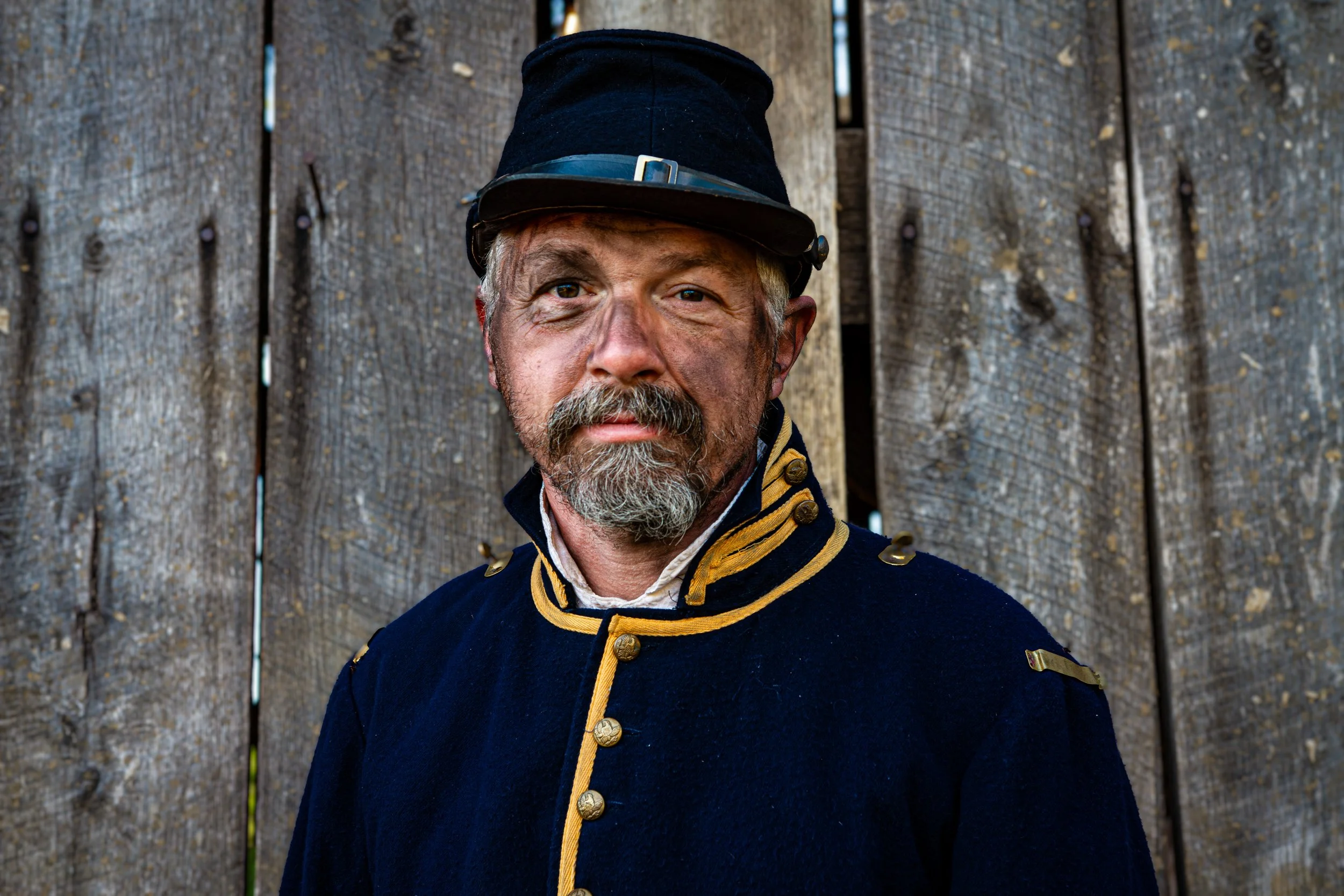 A man dressed in a historical military uniform, including a dark coat with gold buttons and trim, a matching hat with a brim, and a white shirt underneath. The man has a beard and mustache, with a worn face. He is standing in front of a weathered woo