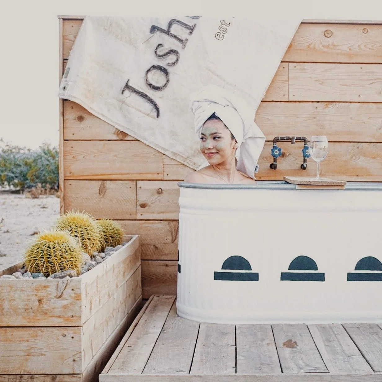 Woman with a towel wrapped around her head relaxing in a vintage bathtub outdoors next to a wooden fence and planter with yellow cacti.