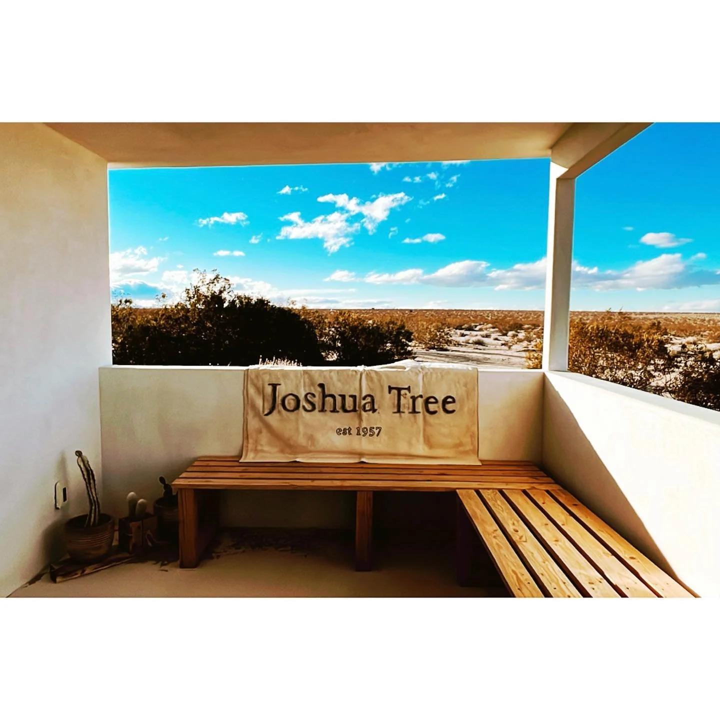 Balcony with wooden bench and sign reading 'Joshua Tree est 1957', overlooking desert landscape with blue sky and clouds.