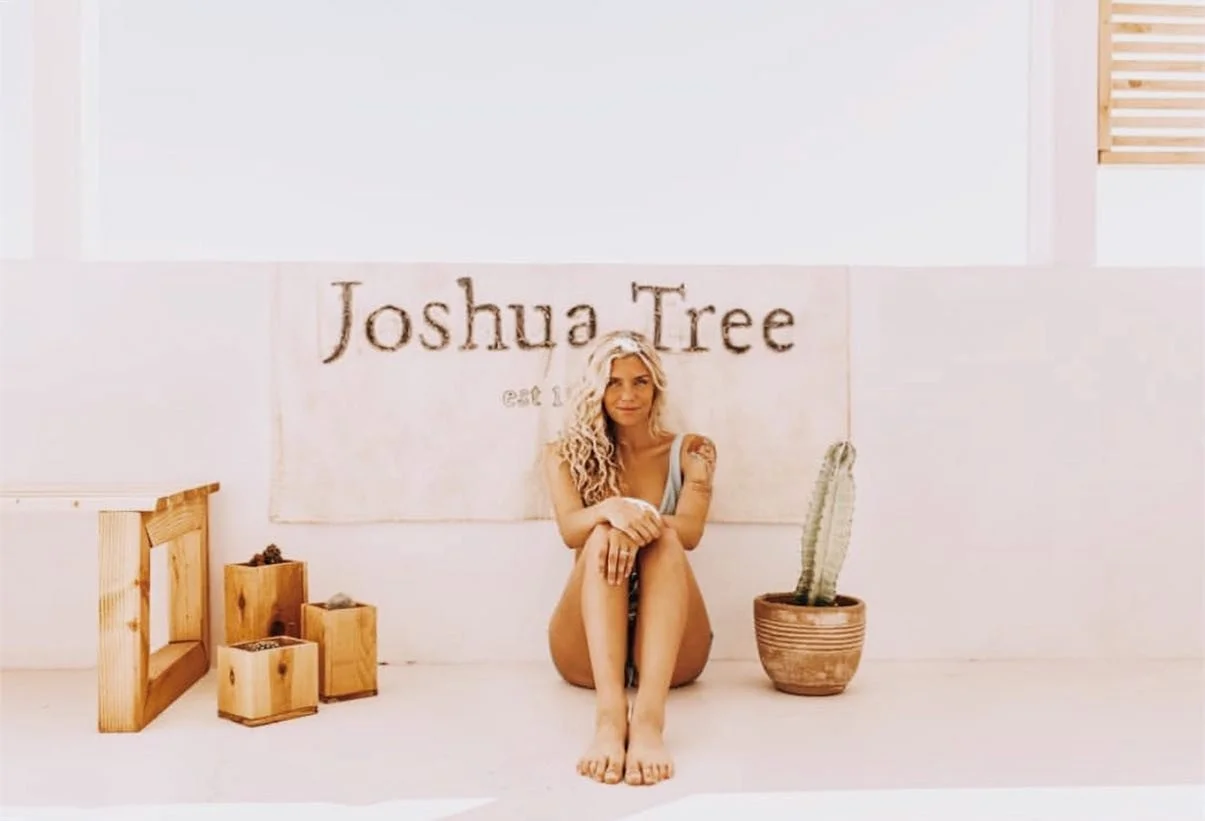 A woman with blonde curly hair sitting on the floor in front of a sign that reads "Joshua Tree" in a bright room with a potted cactus and wooden boxes around her.
