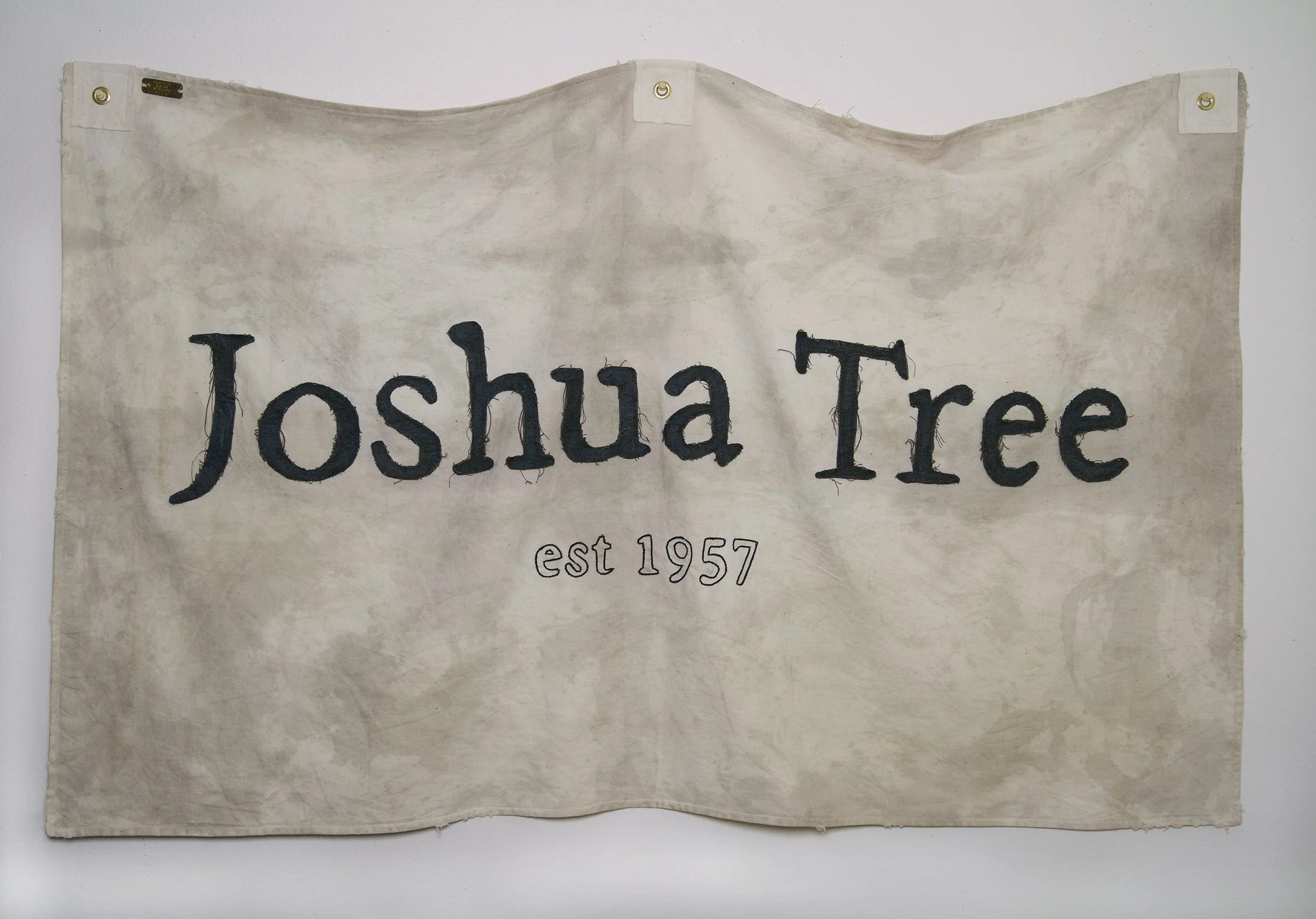 Fabric banner with the words 'Joshua Tree' in large black letters and 'est 1957' below, hung on a white wall.
