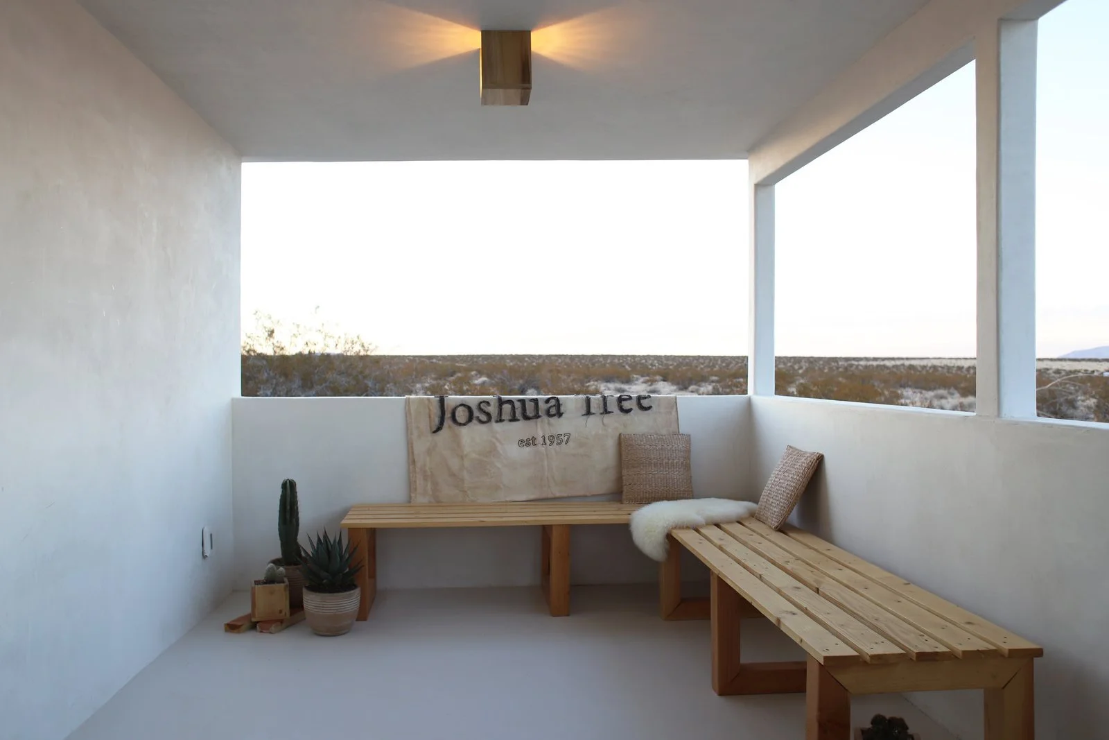 Minimalist balcony with wooden bench and small table, featuring a beige blanket with black text, a pillow, a furry throw, and potted cacti, overlooking a desert landscape.
