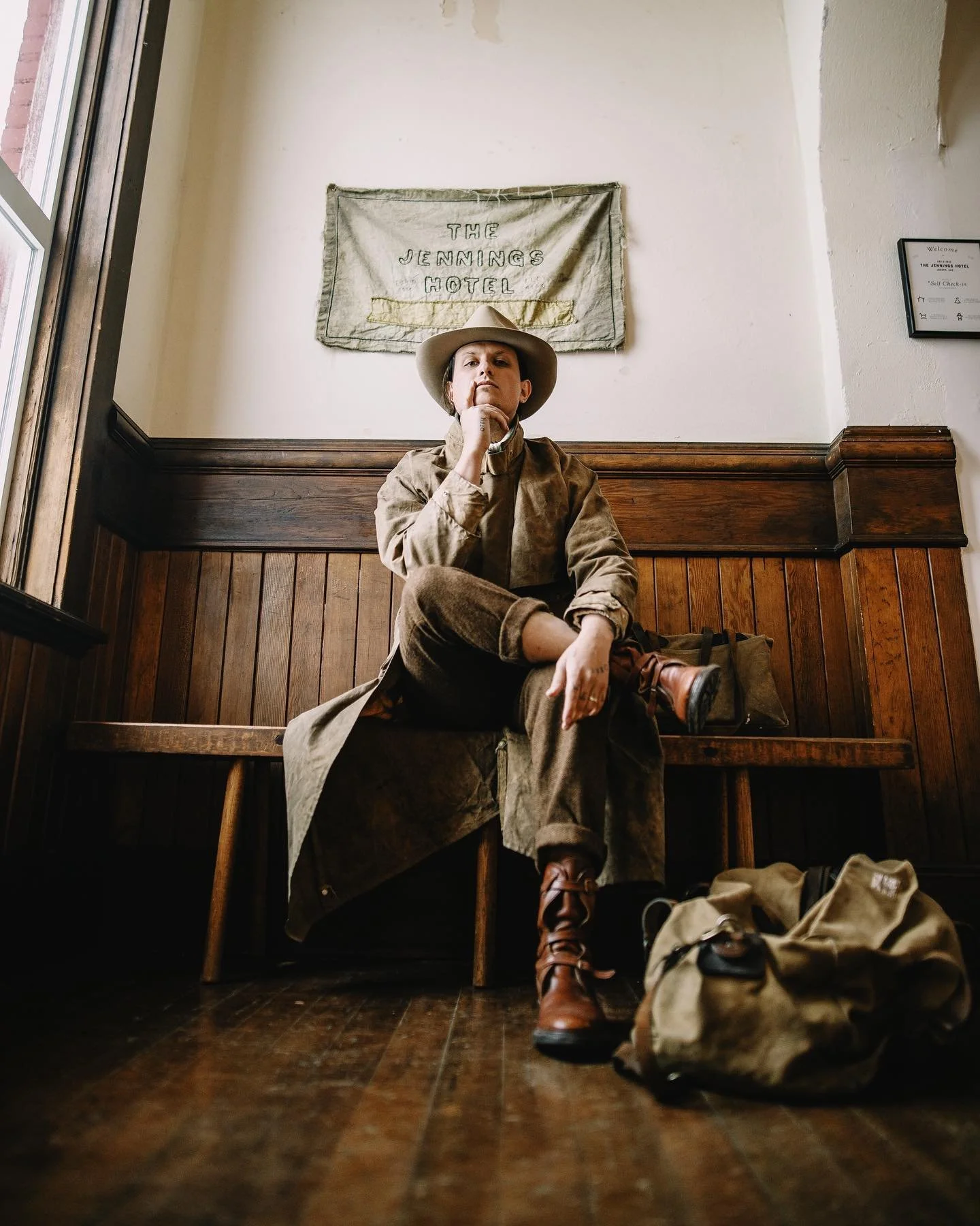 Christian Watson of 1924.us dressed in vintage western attire sitting on a wooden bench in a rustic indoor setting, with a flag reading 'The Jennings Hotel' hanging on the wall behind her.