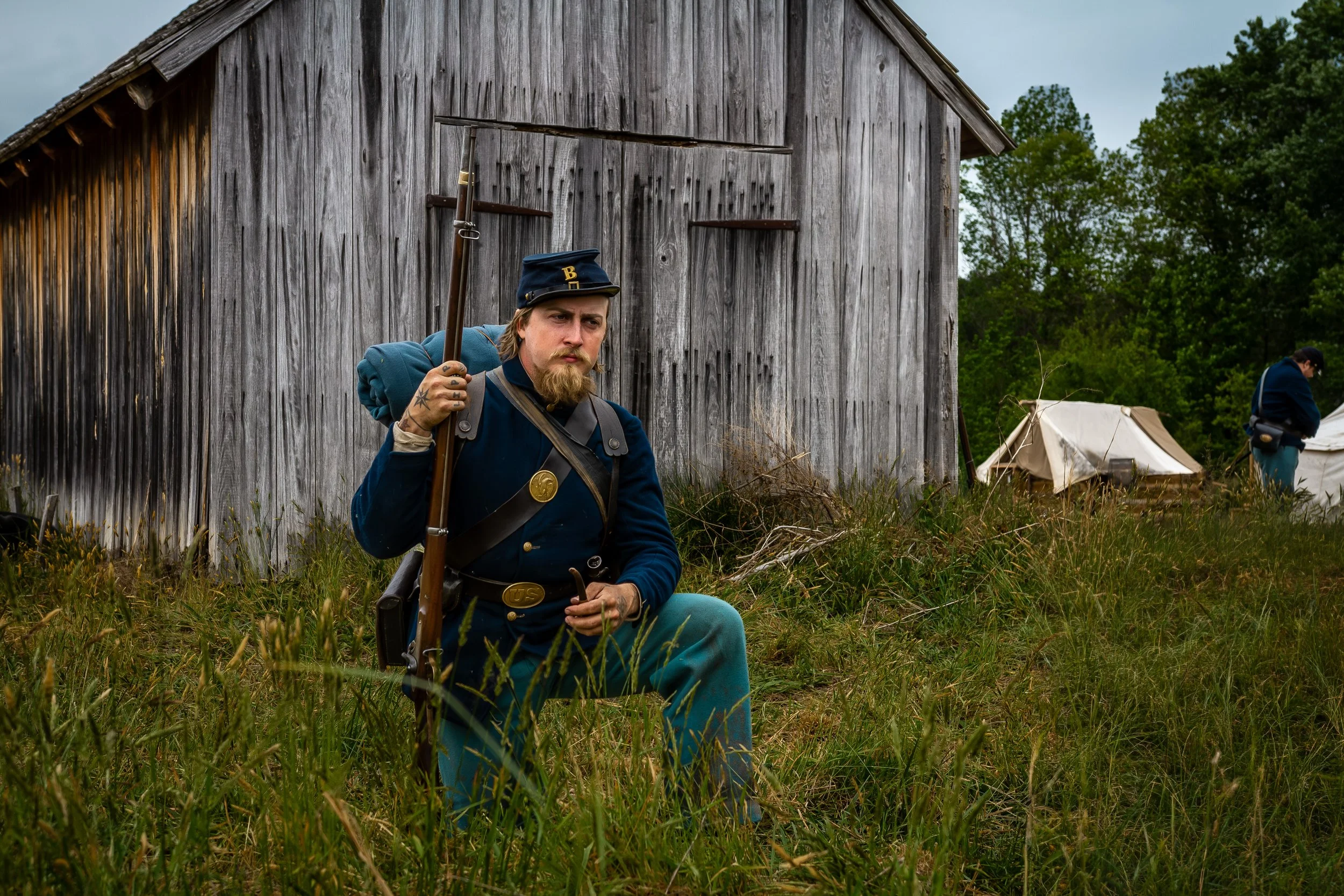 A man dressed in historical military uniform with a blue coat, sitting in tall grass near a weathered wooden barn, holding a long rifle, with tents and another person in historical clothing in the background outdoors.
