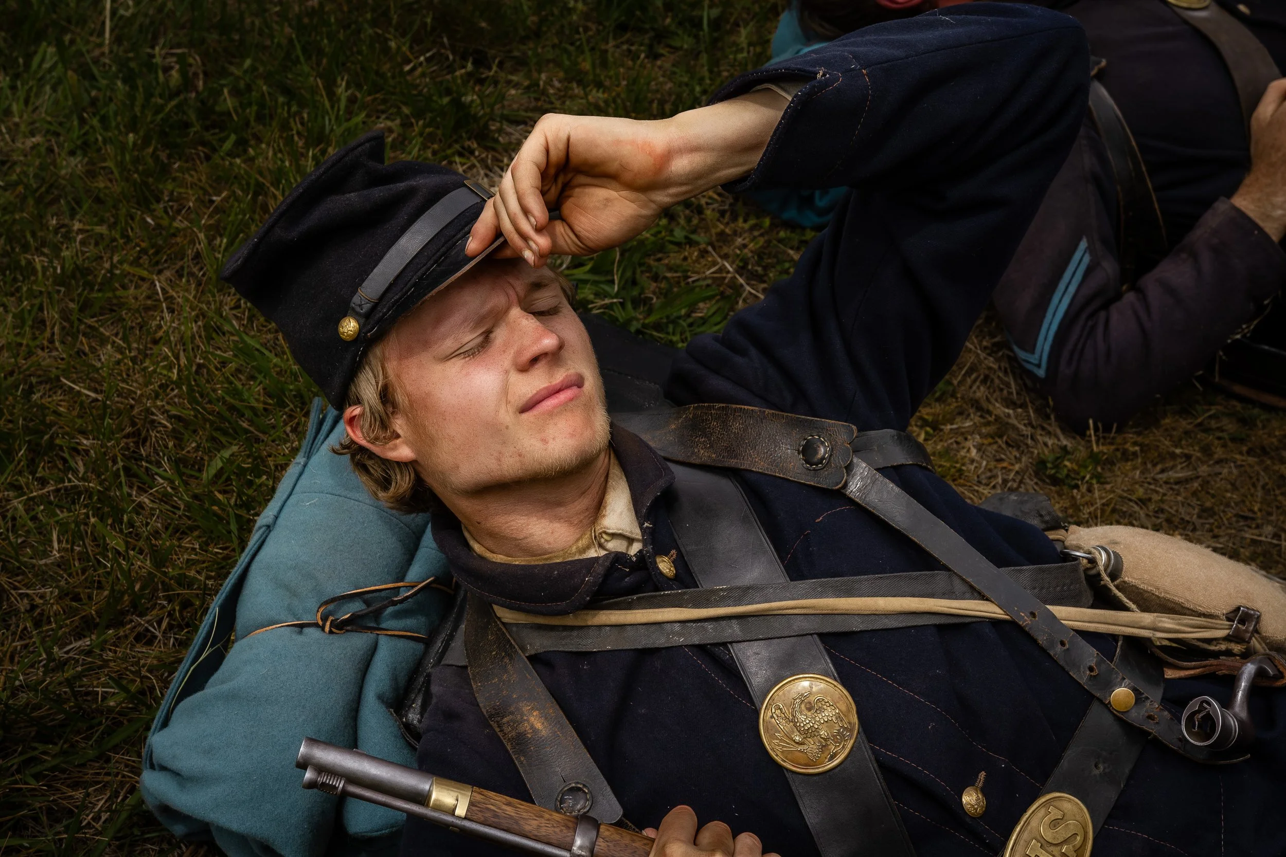 A man dressed as a soldier lying on the ground with eyes closed, wearing a military uniform and hat, holding a rifle, with medals on his chest, in a grassy outdoor setting.