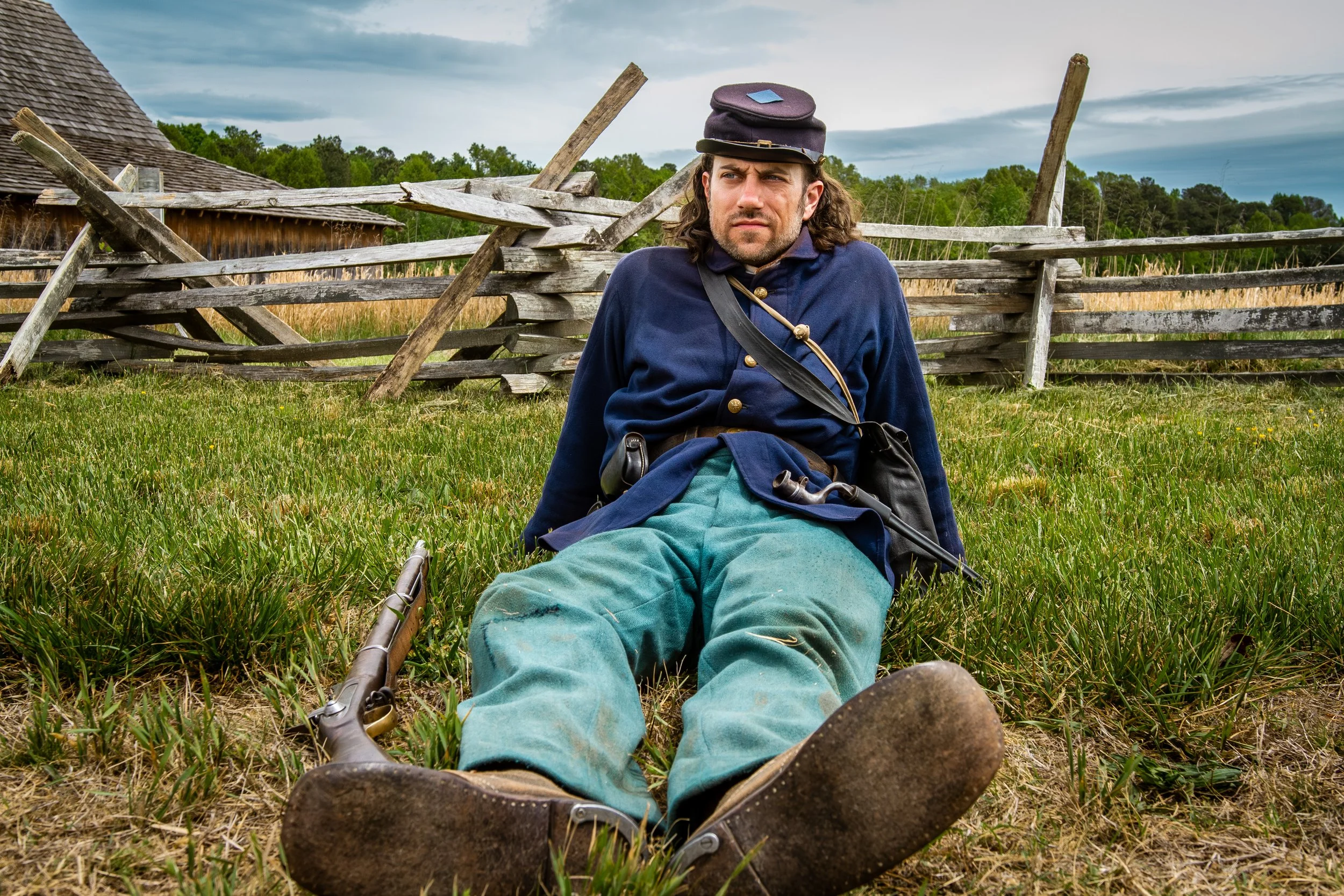 A man dressed in Civil War era clothing sitting on the grass in front of a broken wooden fence, with an old wooden building and green trees in the background, and cloudy sky overhead.