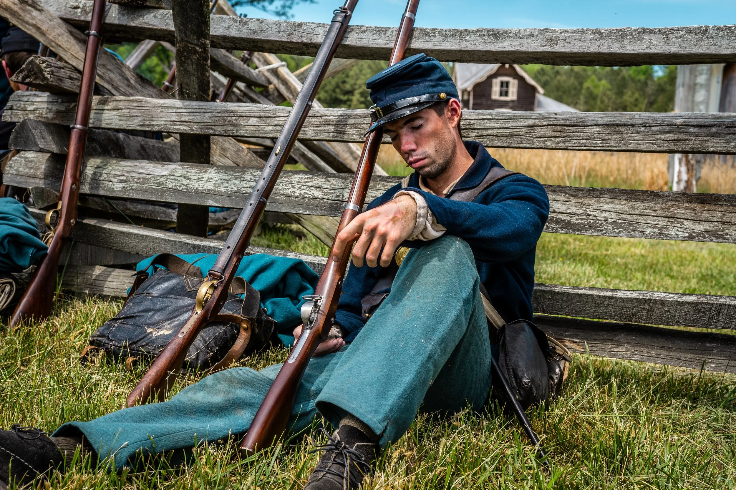 A young soldier in a blue military uniform and hat, sitting on the grass with his back leaning against a wooden fence, looking at a smartphone. There are rifles and gear beside him, with a house and green trees in the background.