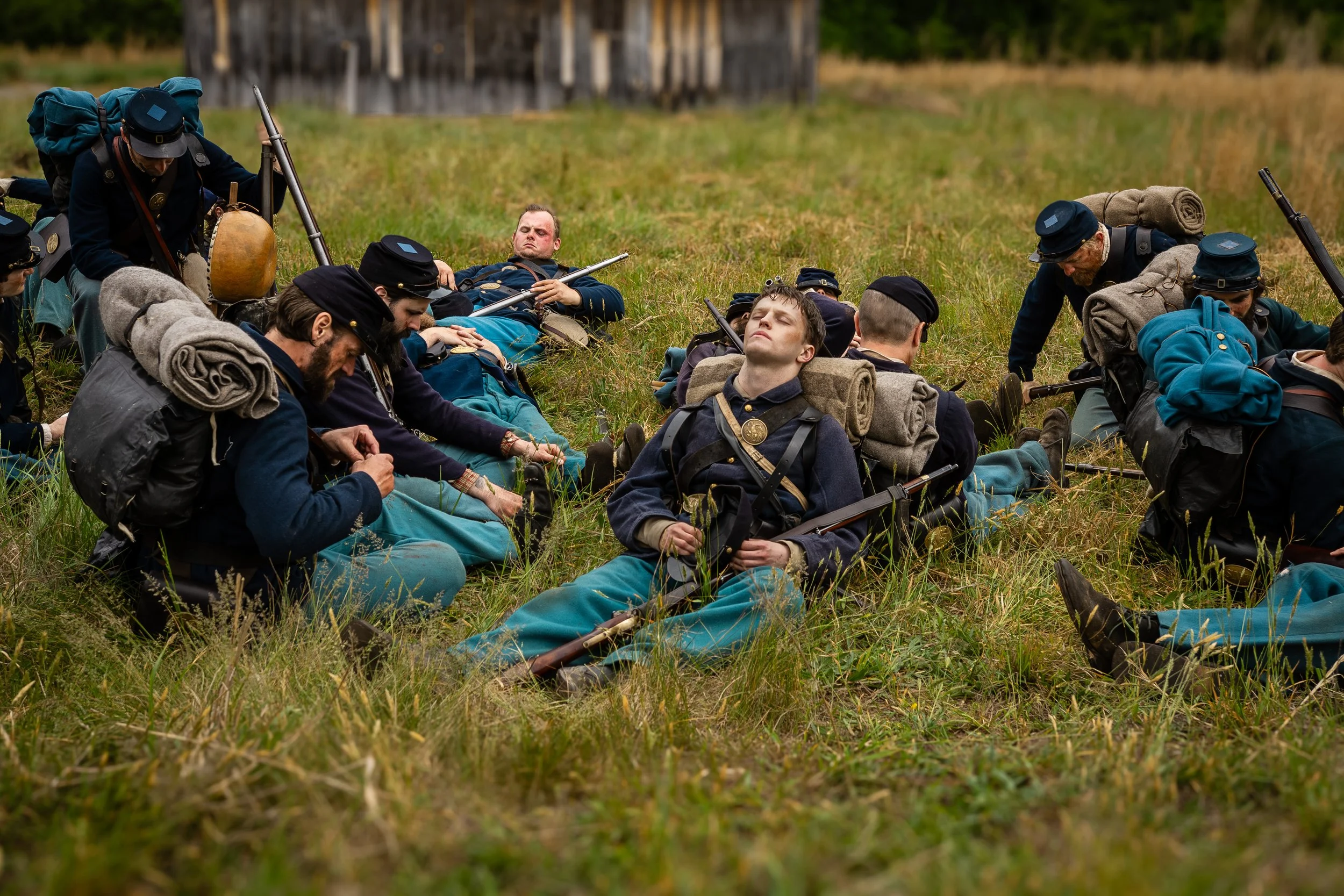 Group of people dressed in historical military uniforms resting on a grassy field in a reenactment scene.