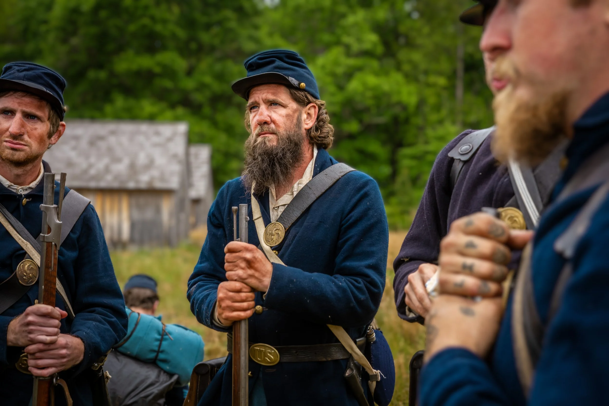 Group of men dressed as Civil War soldiers in uniform, holding rifles, standing outdoors with green trees in the background.