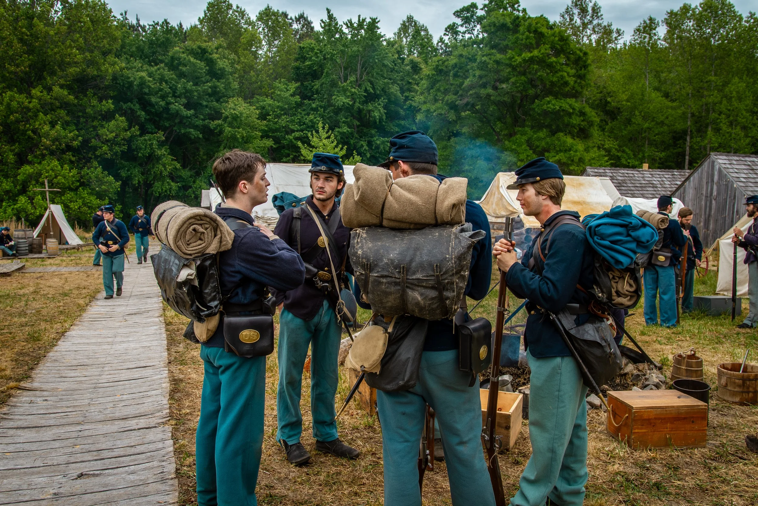Reenactment of a Civil War-era encampment with men dressed in 19th-century military uniforms, carrying gear, and standing on a wooden pathway surrounded by tents and greenery.