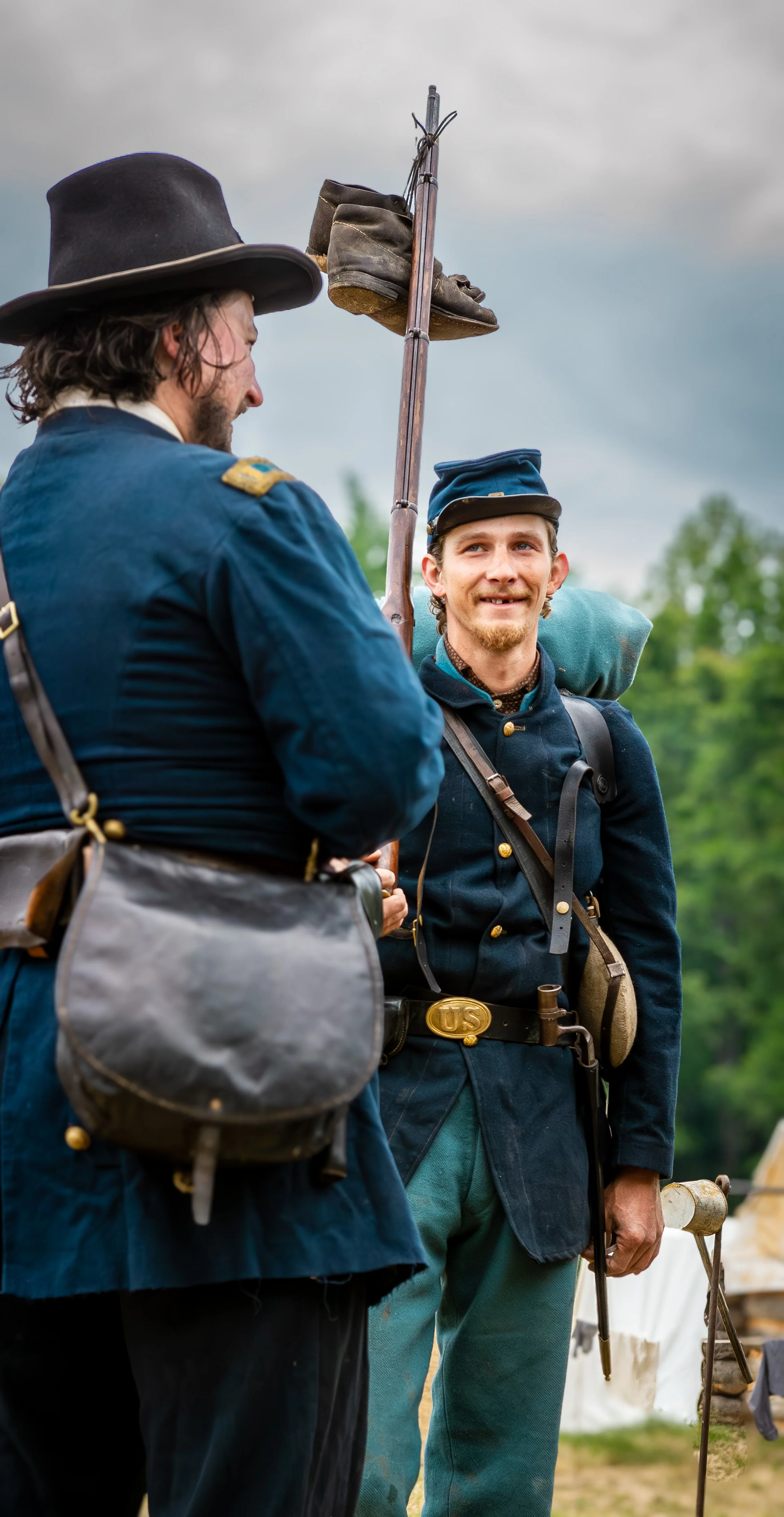 Two men dressed in Civil War era military uniforms, with one man holding a musket, standing outdoors with trees and a cloudy sky in the background.