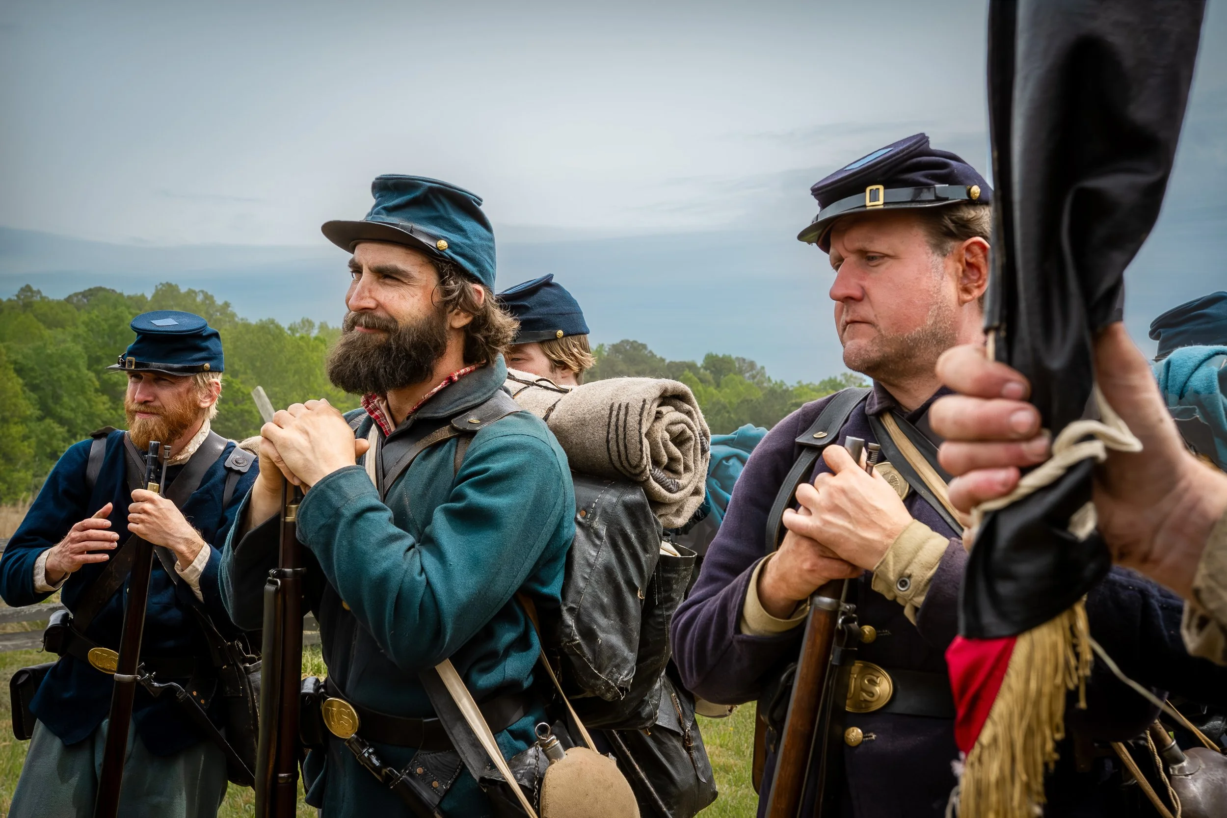 Men dressed as soldiers from the Civil War era, wearing uniforms with blue and dark colors, standing outdoors with a background of trees and a cloudy sky.