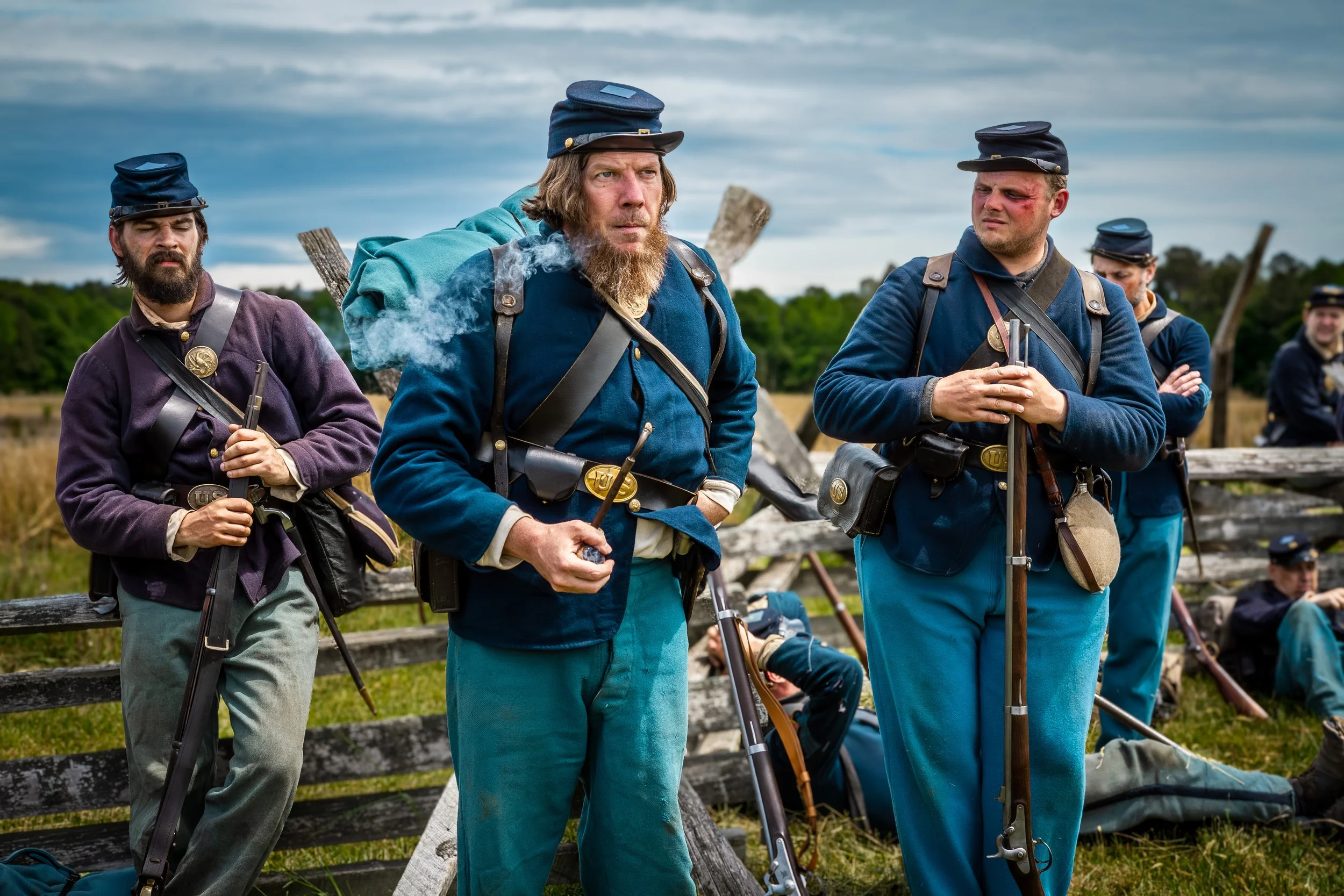 Historical reenactors dressed as 19th-century soldiers in blue and gray uniforms standing outdoors during daytime with a cloudy sky and greenery in the background.
