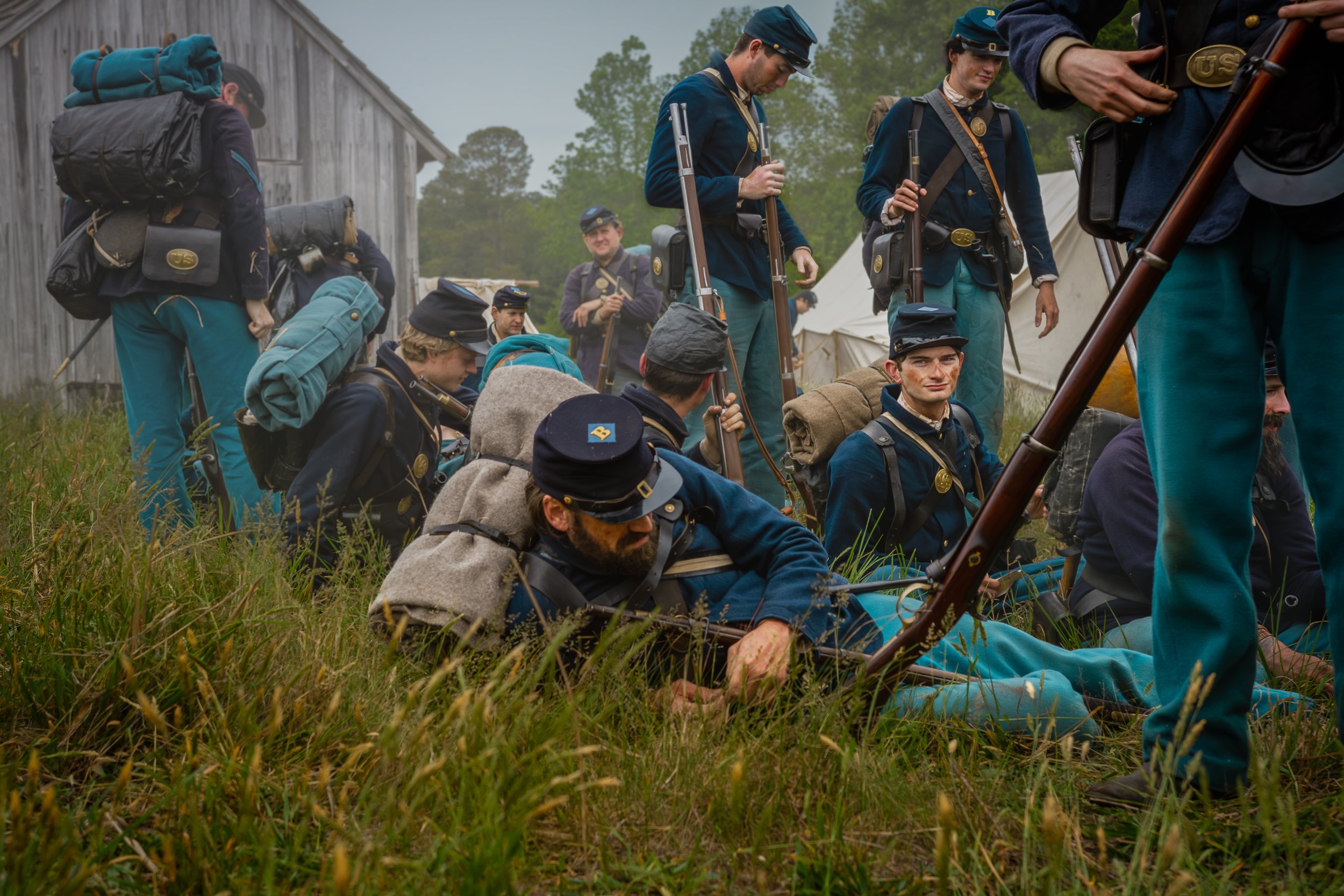 A group of people dressed in Civil War era military uniforms, some sitting on the grass and others standing, outside a wooden building with tents in the background.