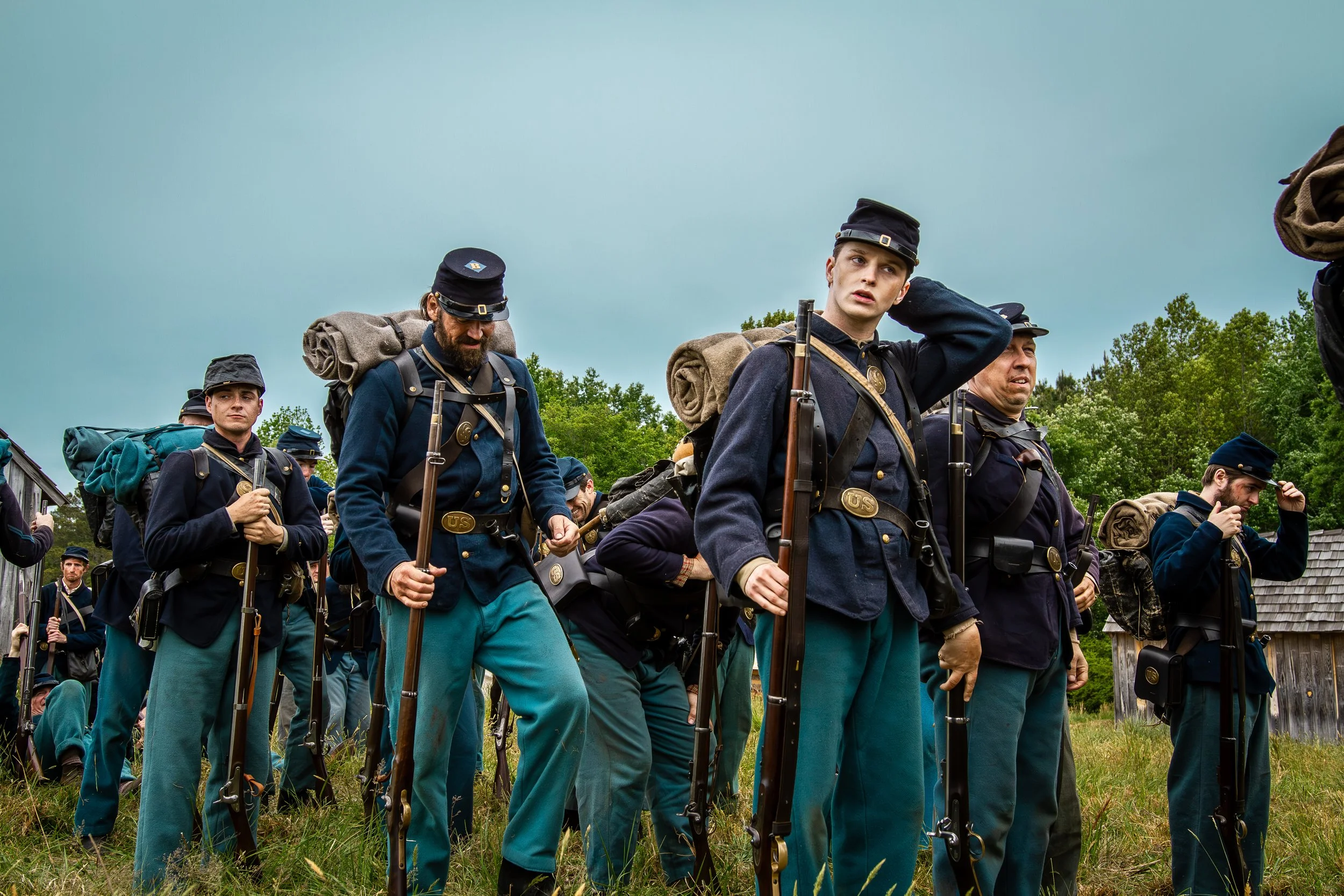 Group of men in Civil War uniforms marching outdoors with backpacks and rifles, with trees and a cloudy sky in the background.