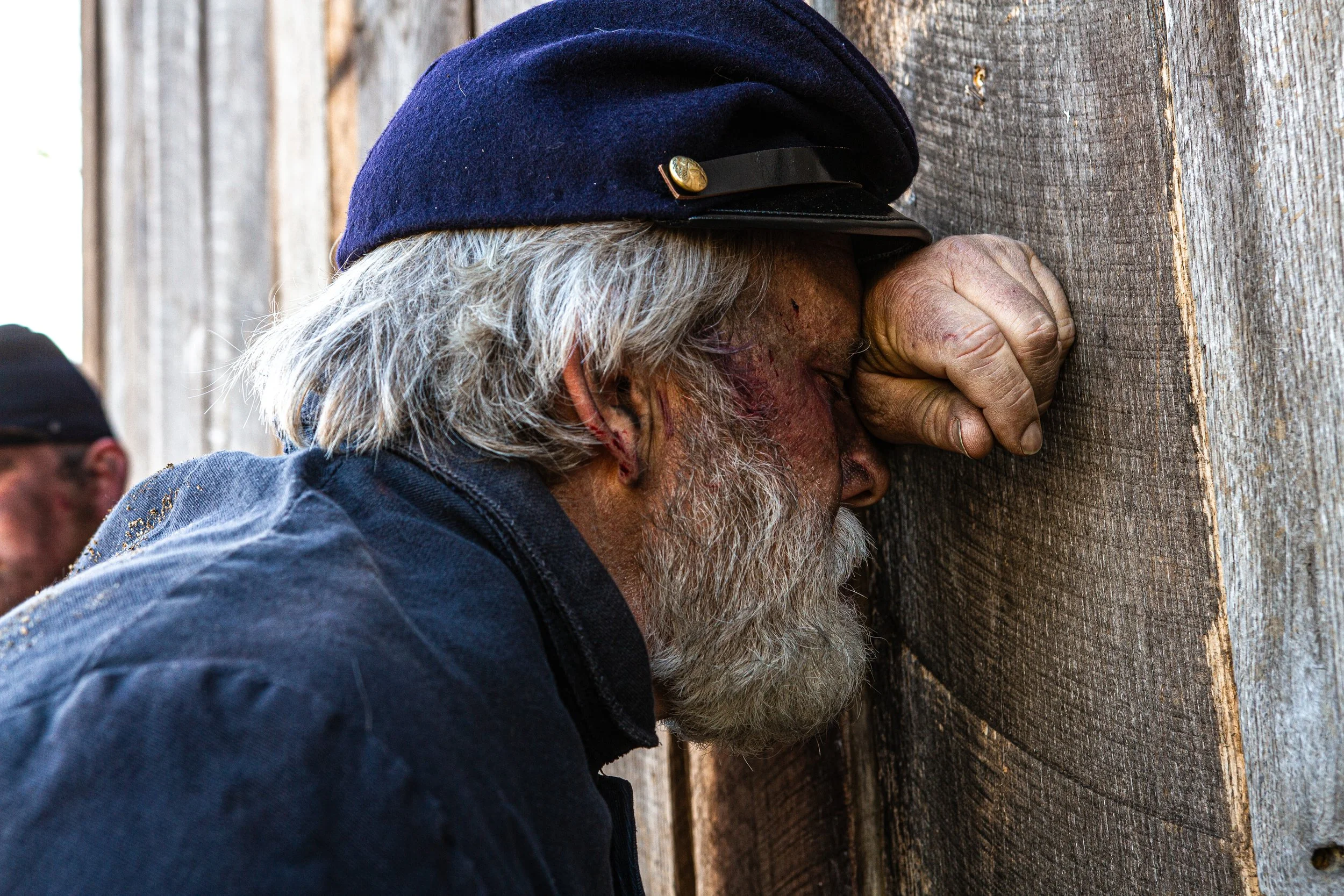 Elderly man with gray hair and beard, wearing a navy blue cap, with eyes closed and forehead pressed against wooden surface.
