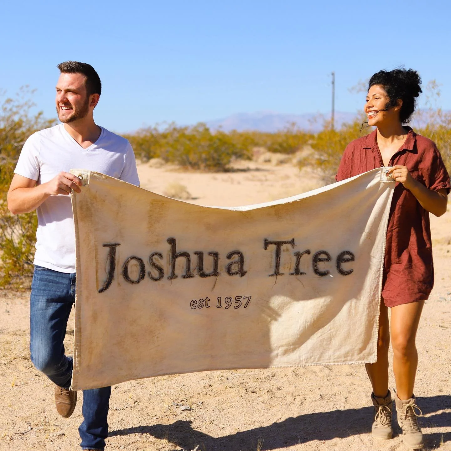 Two people holding a tan banner that reads 'Joshua Tree est 1957' in a desert landscape with sparse bushes and clear blue sky.