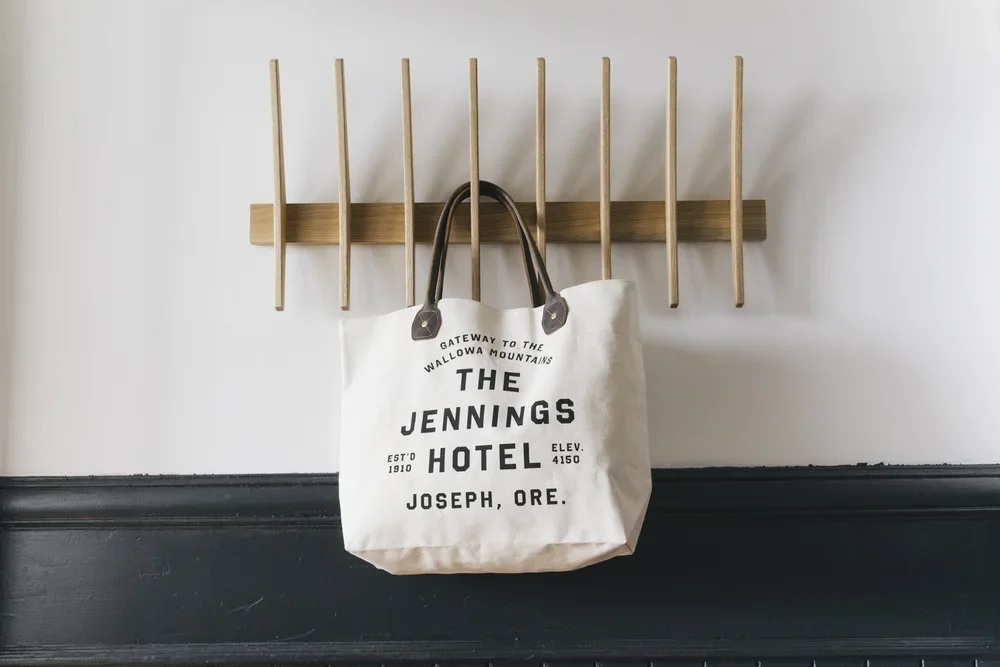 A white tote bag hanging on a wooden wall hook with the text 'The Jennings Hotel, Joseph, Ore.' printed on it.