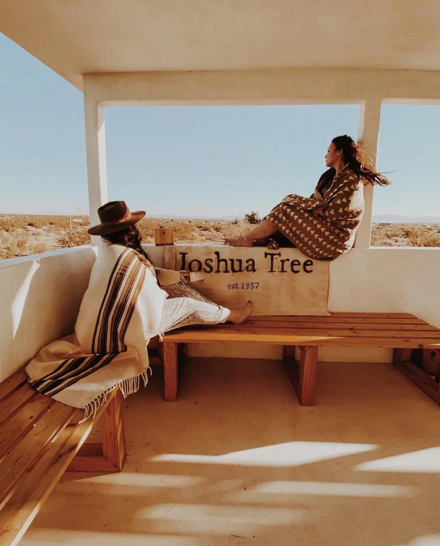 Two women sitting and lounging inside a small, open structure, with one on the bench and the other sitting on the wall, overlooking a desert landscape. The structure has a sign that reads 'Joshua Tree est 1957'.