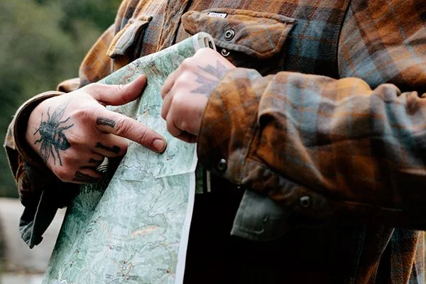 Person holding a folded map outdoors, wearing a plaid jacket with tattoos on their hand.