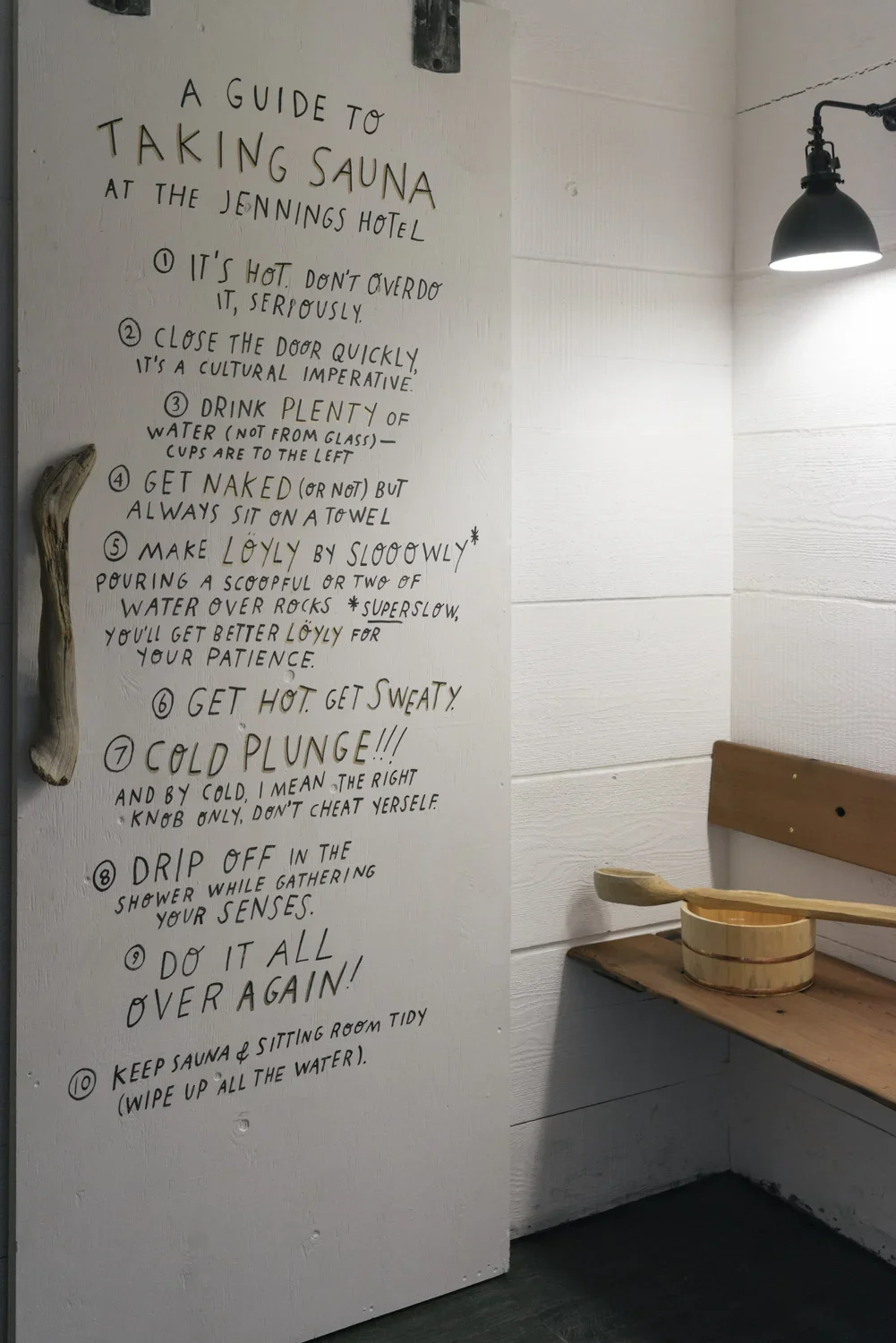 Handwritten guide on a white wall titled 'A Guide to Taking Sauna at the Jennings Hotel.' It lists ten tips with humorous advice for sauna use, including: 1. It's hot, don't overdo it seriously. 2. Close the door quickly, it's a cultural imperative. 
