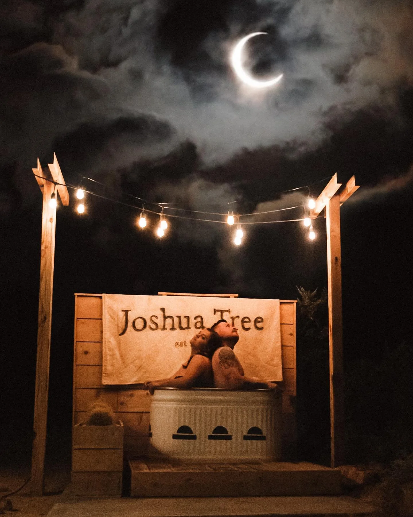 Person in a hot tub at night, illuminated by string lights, with a sign reading 'Joshua Tree' behind them and a crescent moon in the cloudy sky above.