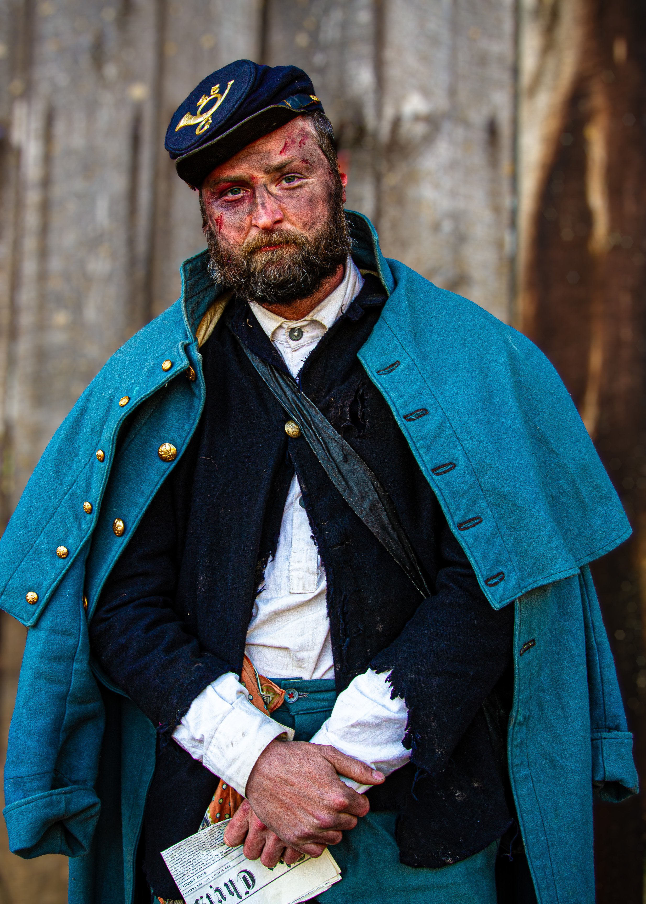 A man dressed as a Civil War soldier with a bloodied and bruised face, wearing a blue coat and hat, holding a newspaper, standing outdoors with a wooden fence in the background.
