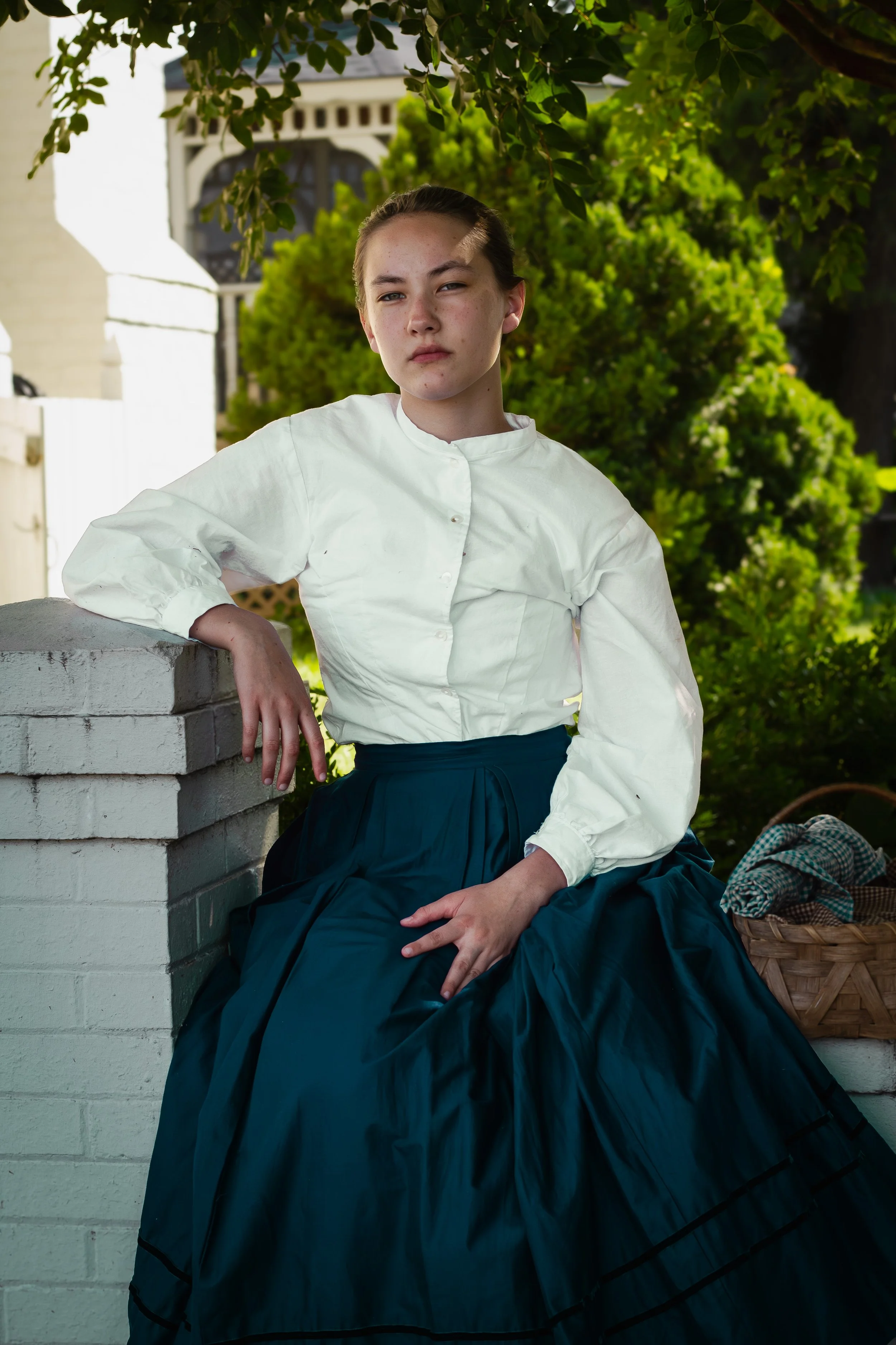 Young woman with dark hair, wearing a white blouse and a dark skirt, sitting outdoors near a white brick wall, with green trees and a house in the background.