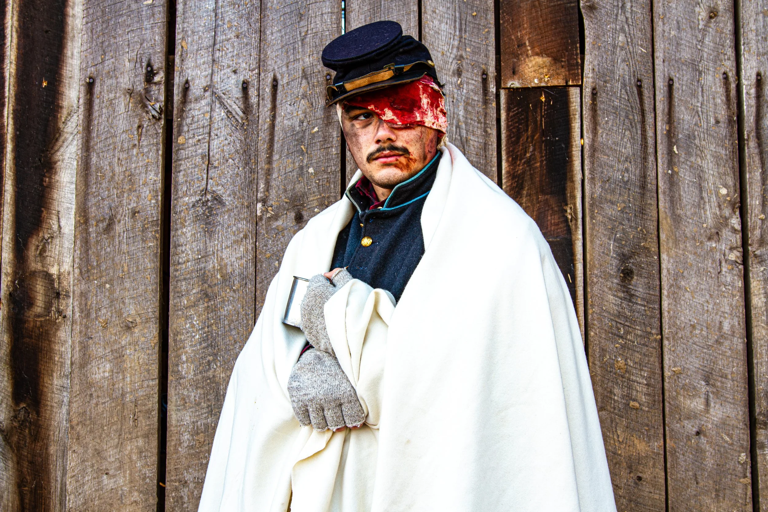A young man with a bloodstained bandage over his right eye, wearing a police officer's hat at an angle, with dirt and blood on his face, standing against a wooden fence background.